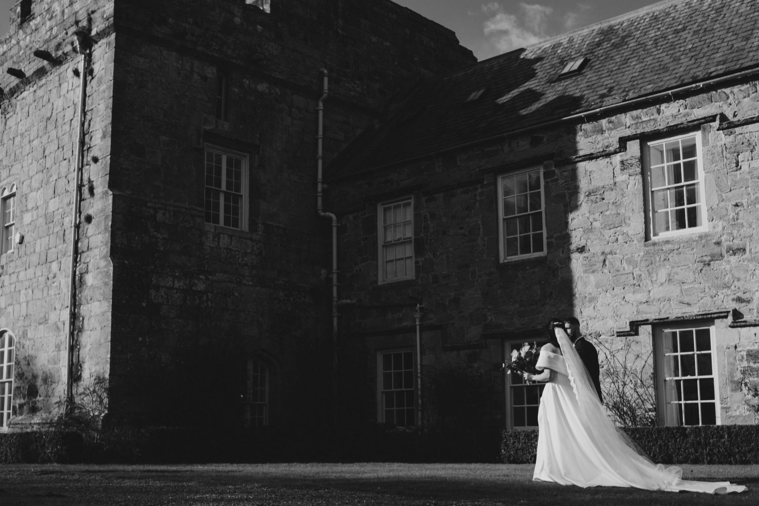 Black and white wedding photo of bride in flowing gown holding bouquet outside historic stone castle venue.