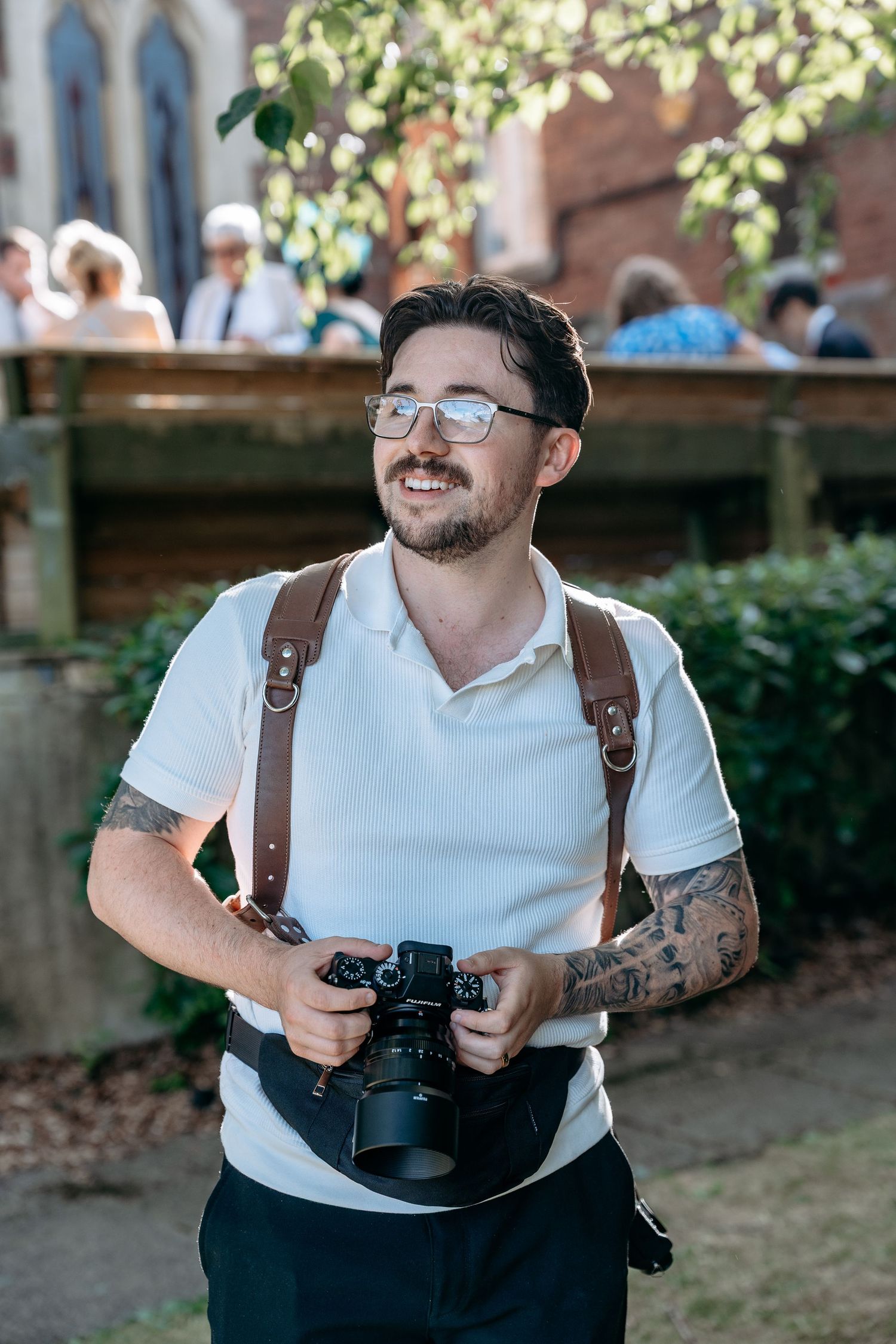 Smiling tattooed photographer holding a camera outdoors at a sunny event, wearing glasses and a white polo shirt.