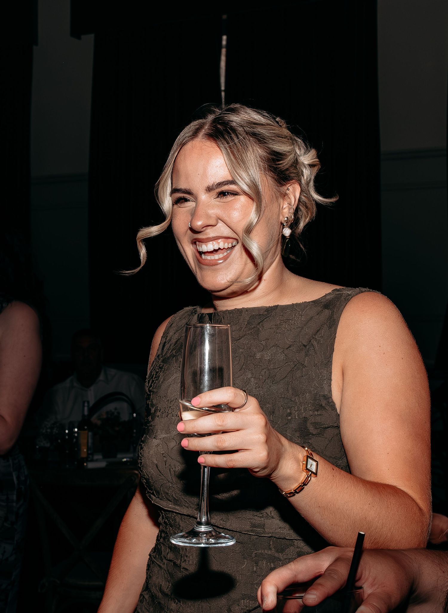 A smiling woman in a brown dress holds a champagne flute at an evening event, wearing pearl earrings and a gold watch.
