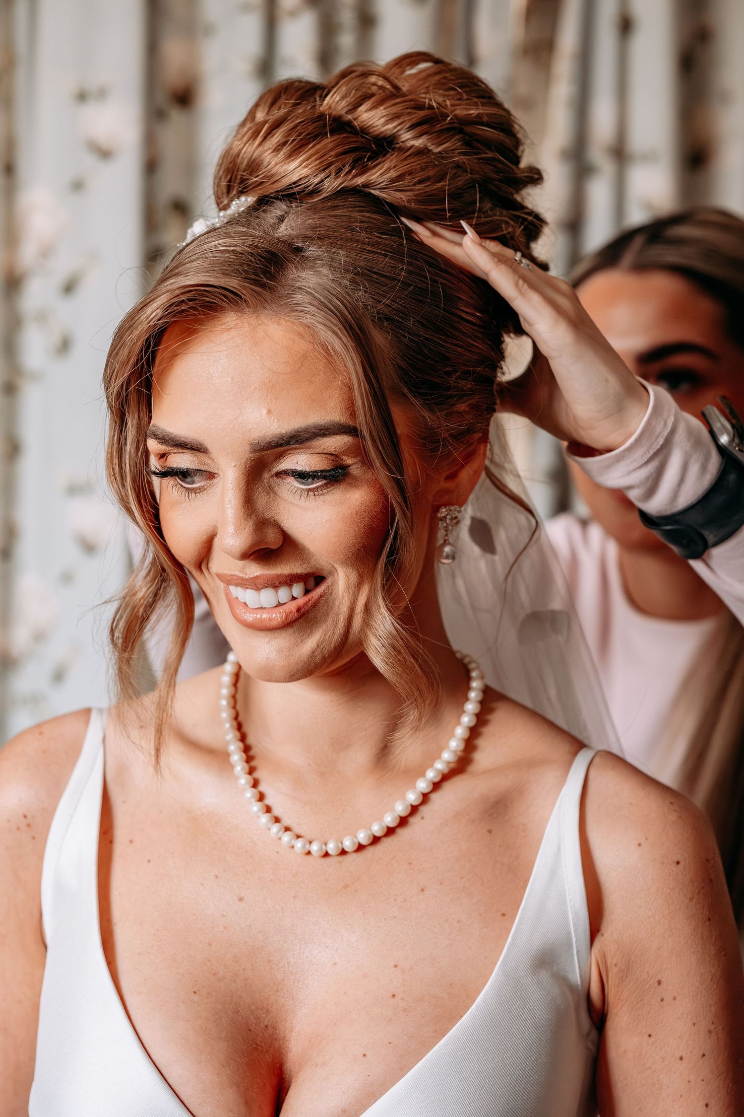 Smiling bride in pearl necklace gets braided updo styled by hairdresser on wedding day.