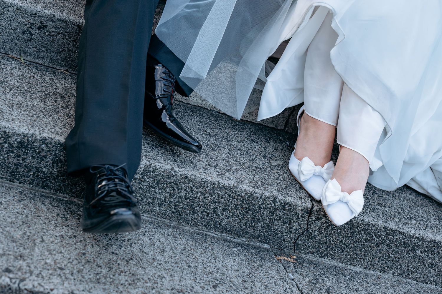 Bride and groom sitting on stone steps, showing black dress shoes and white bridal heels with bow details.