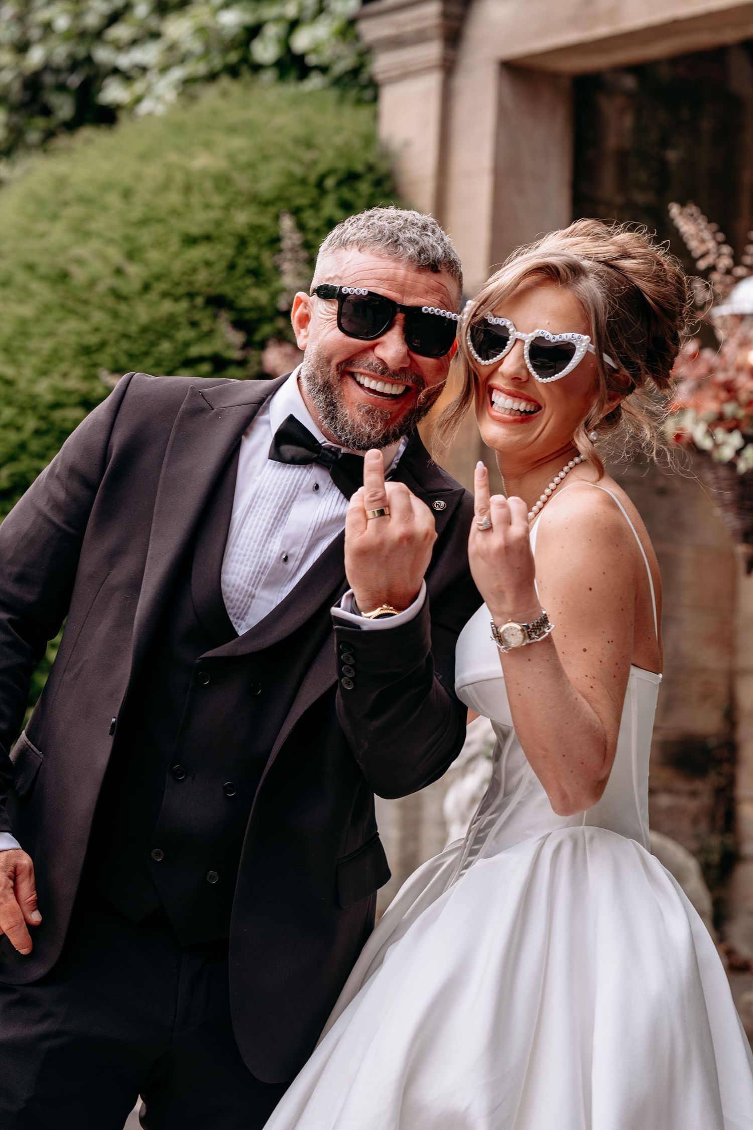 Laughing bride and groom in sunglasses showing wedding rings, wearing white gown and dark tuxedo outdoors.