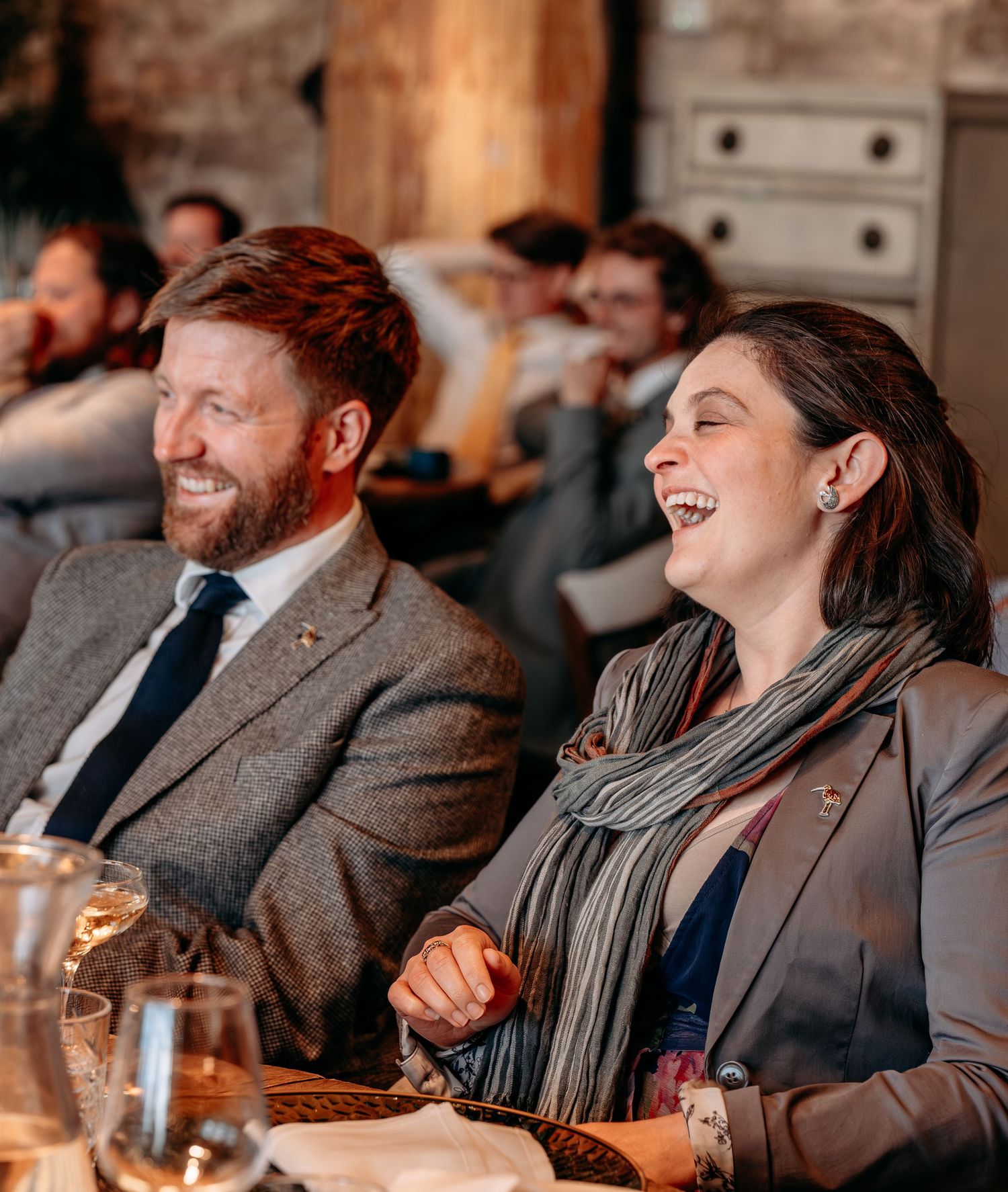 A man and woman laugh joyfully at an indoor event, dressed in smart casual attire with drinks on the table.
