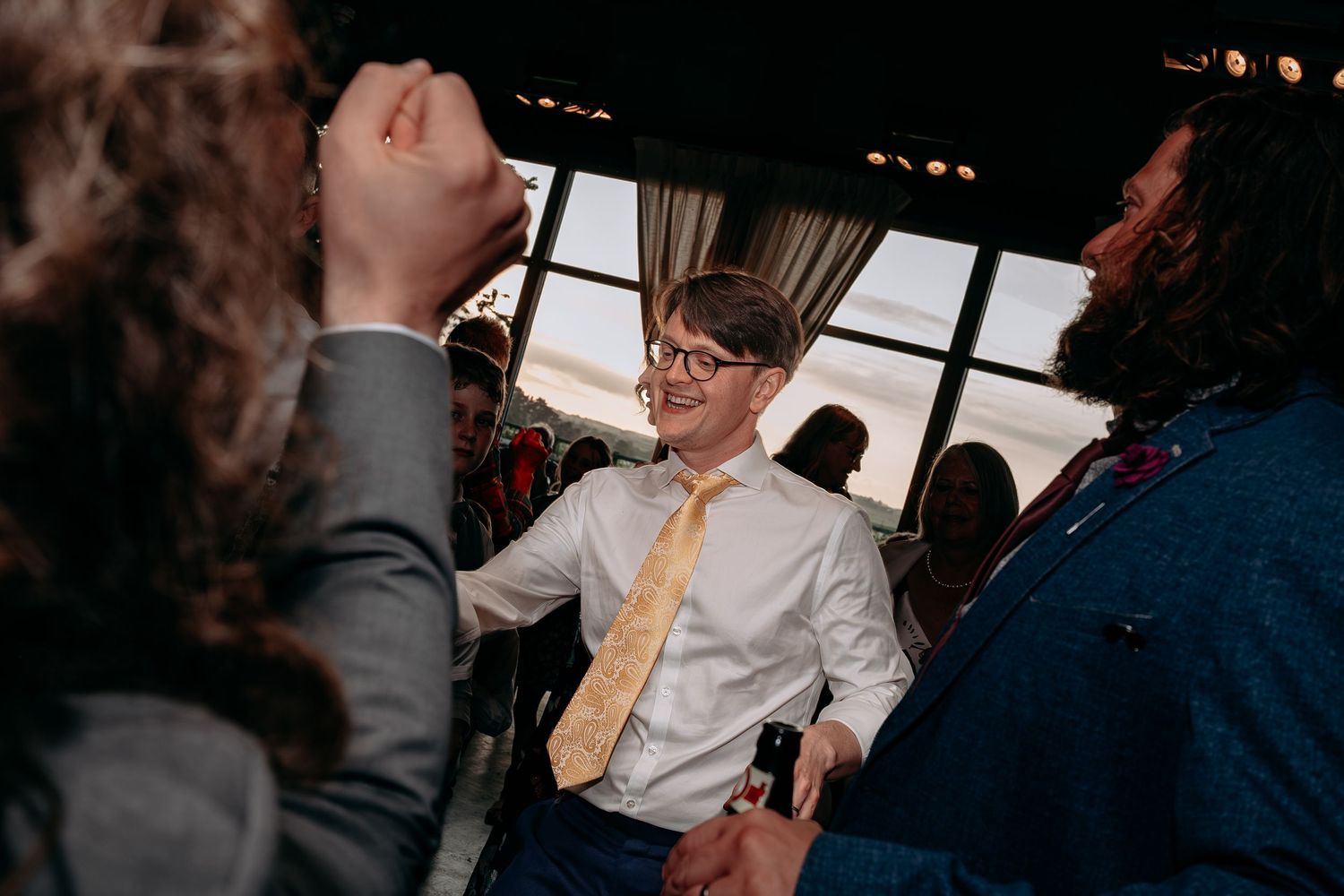 Young boy in glasses and gold tie smiling at wedding reception celebration indoors.