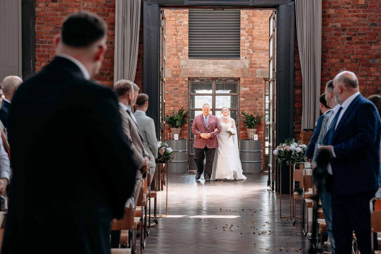 Bride walks down the aisle with escort in a rustic brick wedding venue, guests seated on wooden pews.