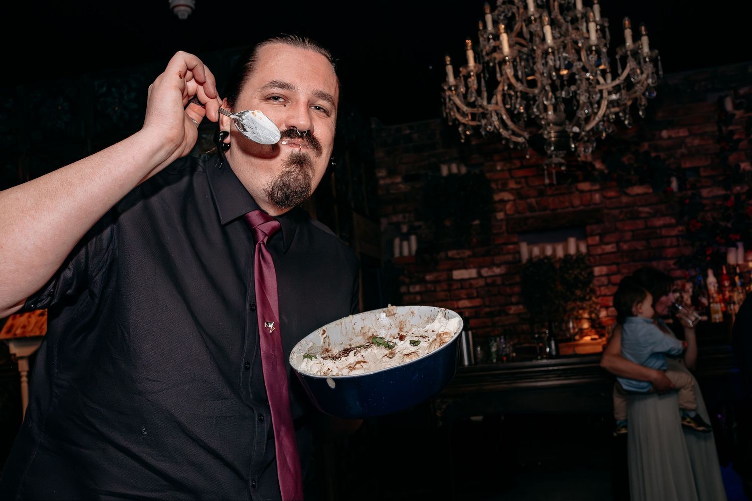 Man in black shirt holding blue bowl of dessert, smiling with spoon, elegant chandelier venue in background.