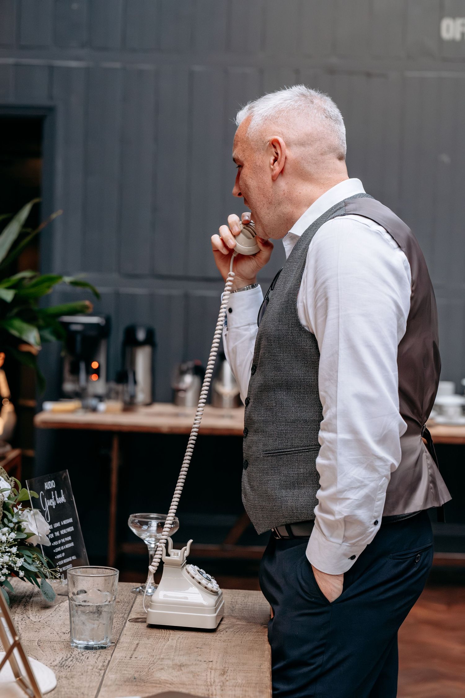 Older man in gray vest speaking into vintage rotary phone handset at wooden table during outdoor event.