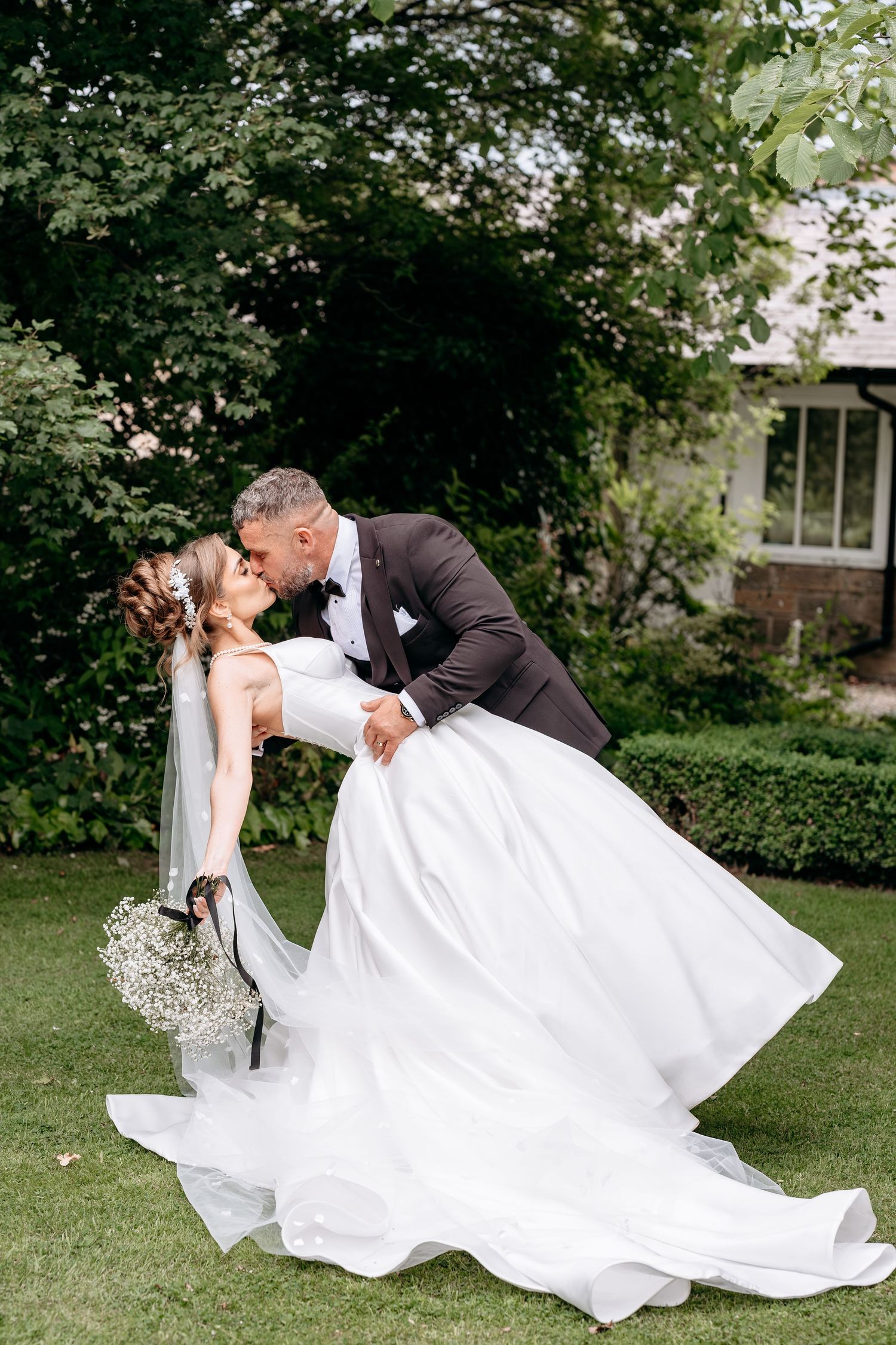 Groom dips bride for a romantic kiss outdoors on green lawn, bride in flowing white gown holding bouquet.