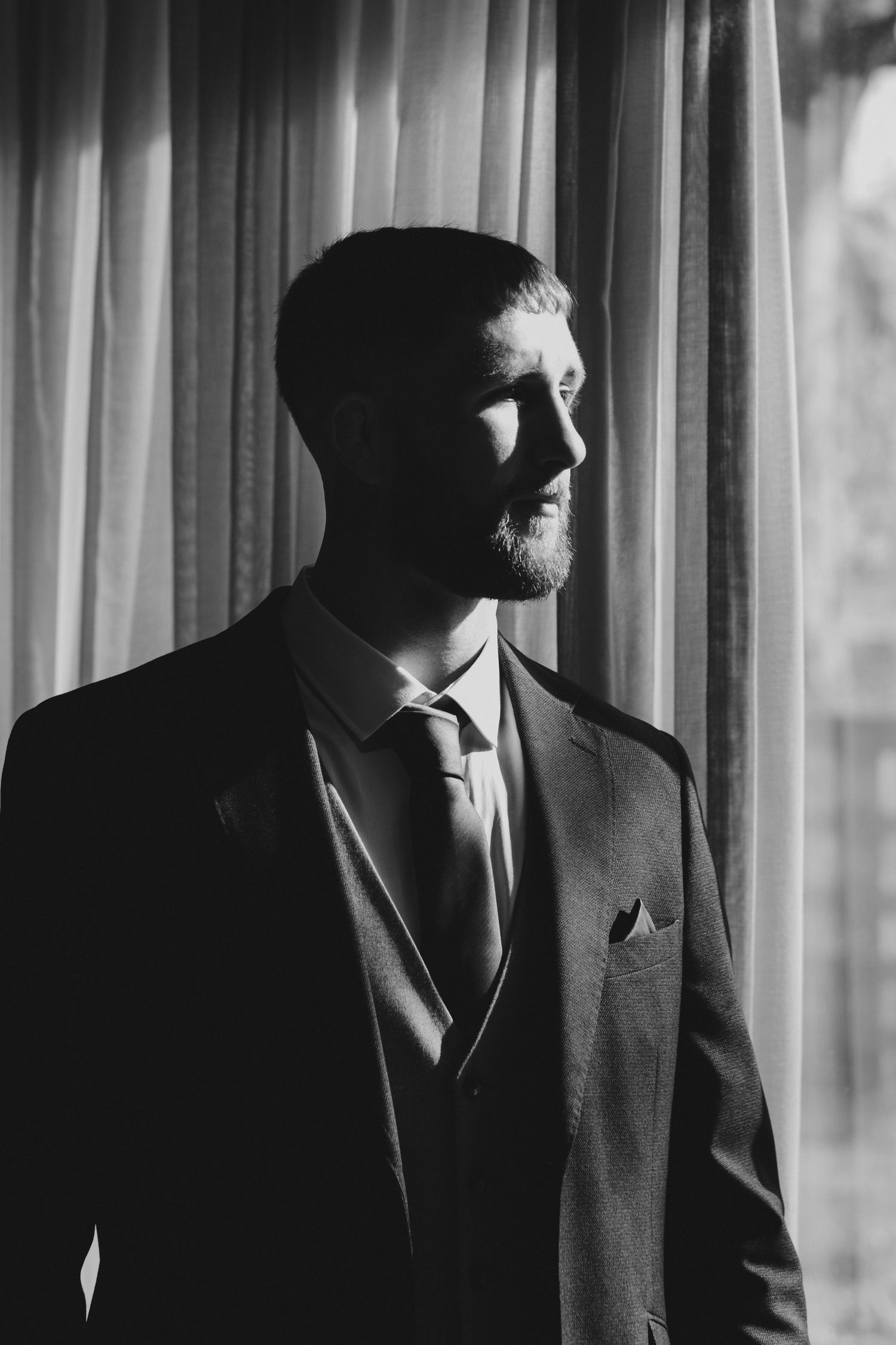 Black and white portrait of a bearded man in a suit and tie standing by a curtained window.