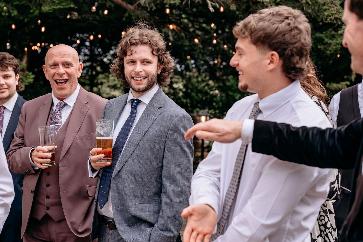 Group of men in suits laughing and socializing outdoors at a wedding reception, holding drinks.