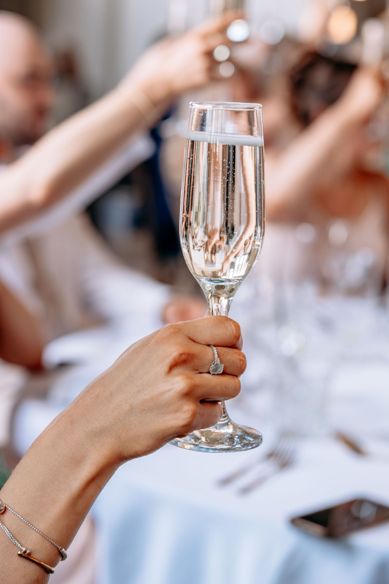 A hand wearing a diamond ring holds a crystal champagne flute at an elegant wedding reception.