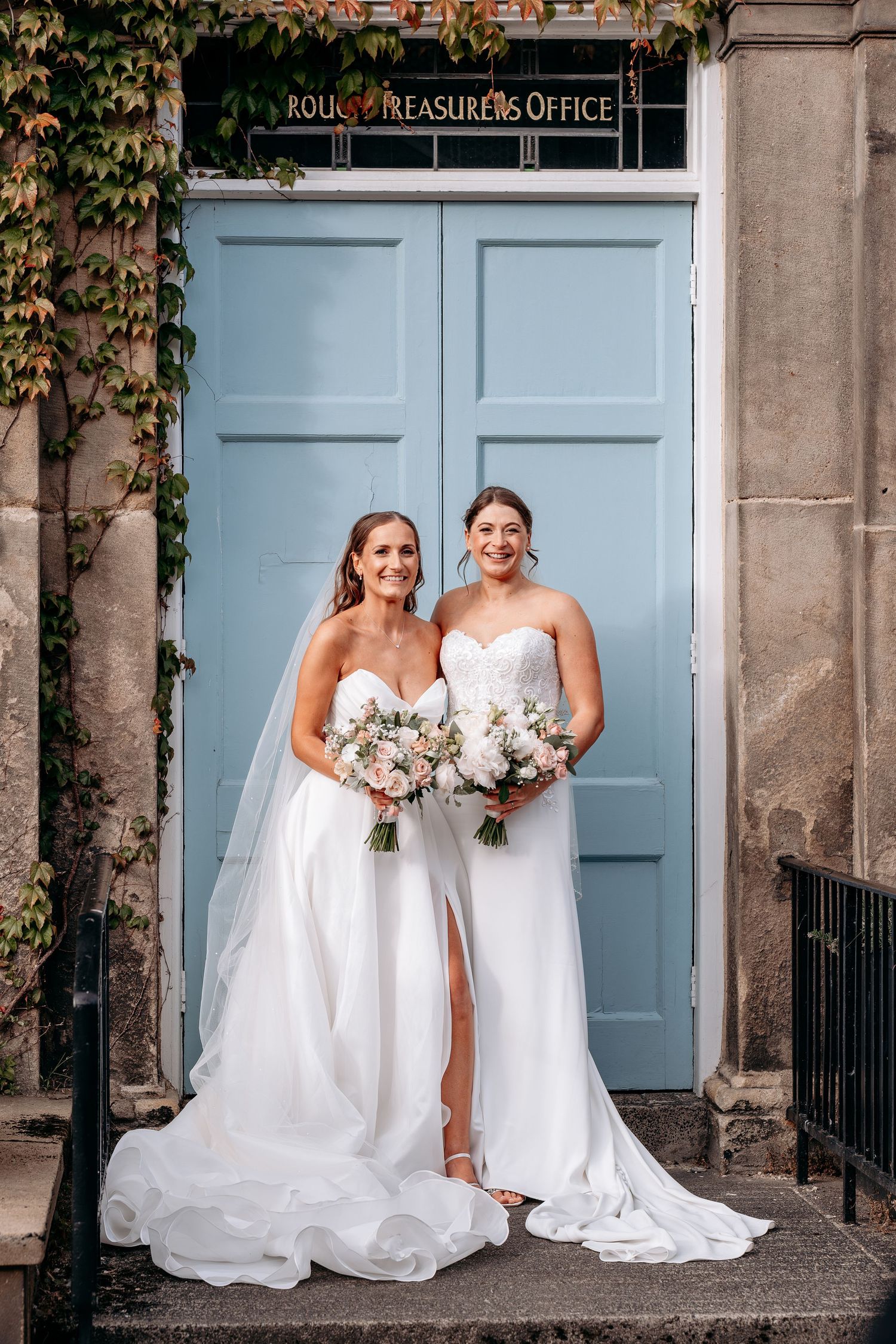 Two brides in white strapless gowns holding floral bouquets pose before a blue door with ivy-covered stone walls.