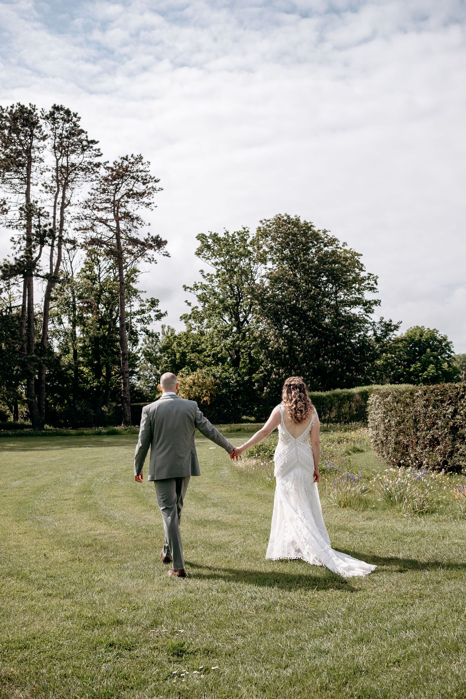 Bride and groom walking hand in hand across lush green lawn surrounded by trees on their wedding day.