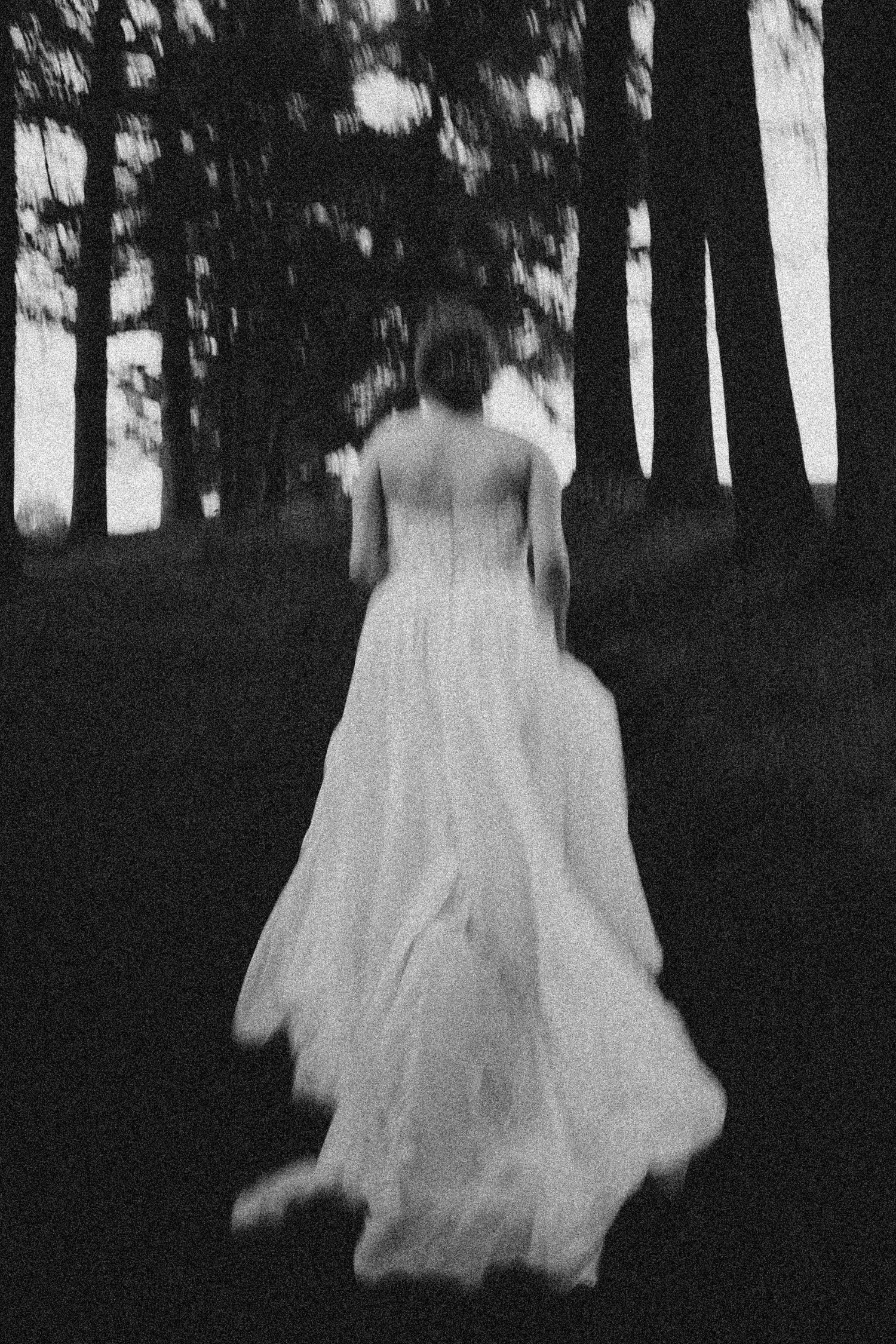 Artistic blurred black and white photo of a bride in a flowing gown among tall trees.