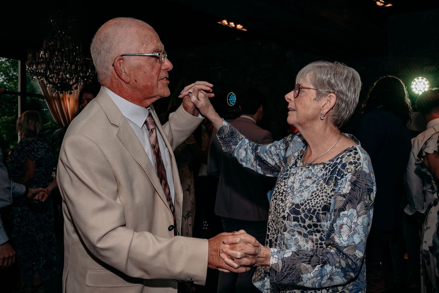 Elderly couple dancing together at a formal event, holding hands and smiling in warm ambient lighting.