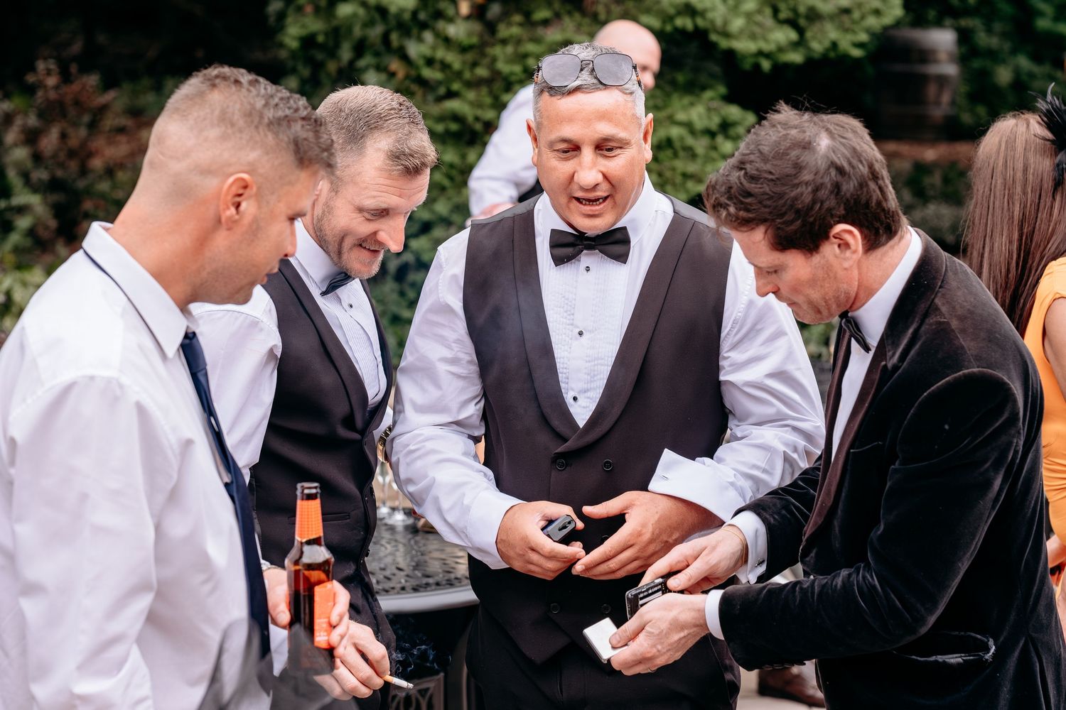 Men in formal attire gather outdoors, examining items, one holding a beer bottle at a wedding reception.