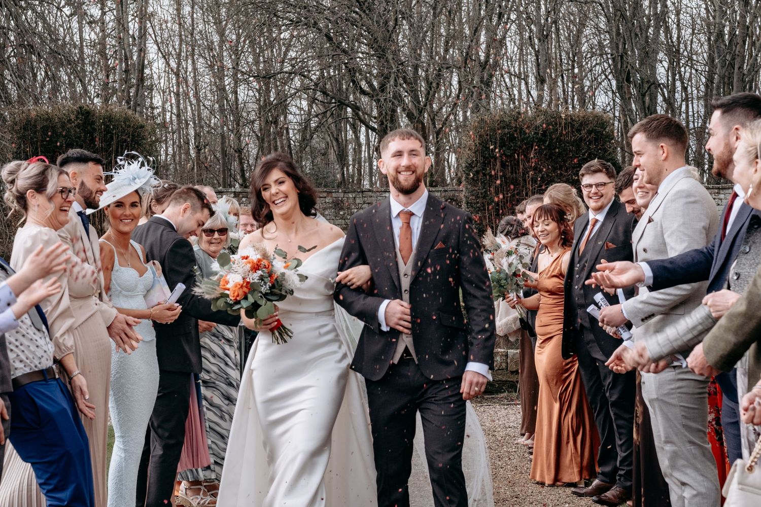 Bride and groom walk through confetti shower with wedding guests cheering in a tree-lined outdoor ceremony.