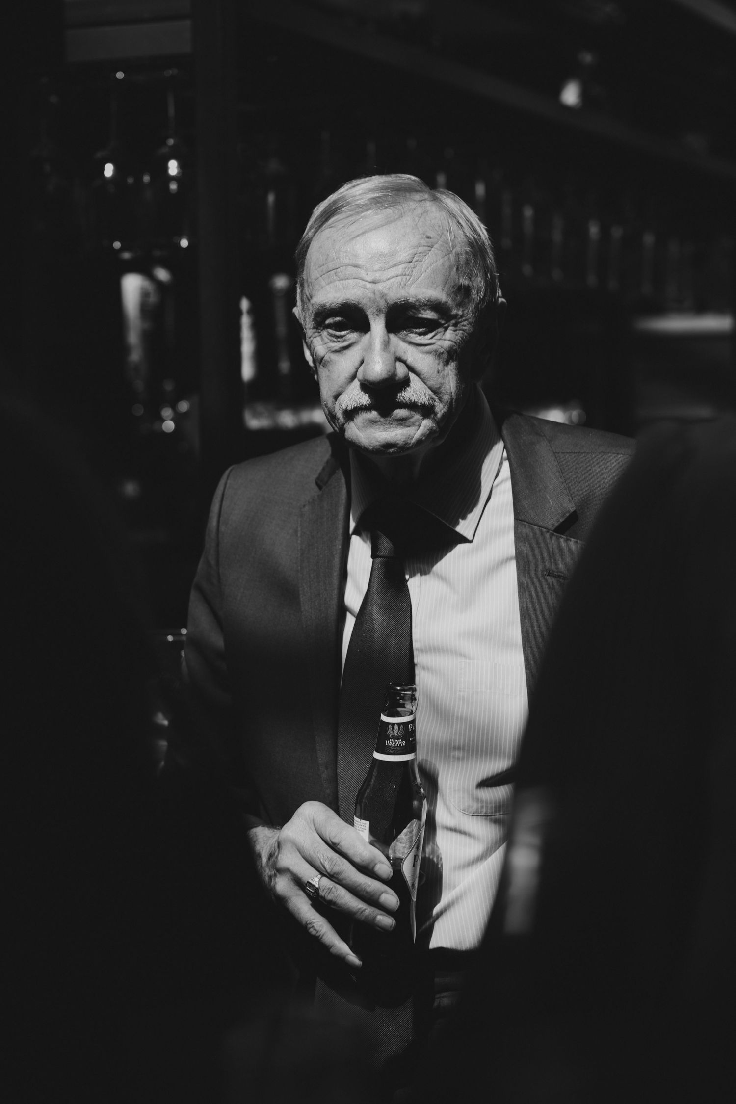 Elderly man in suit and tie holding beer bottle at dimly lit bar, black and white portrait photography.