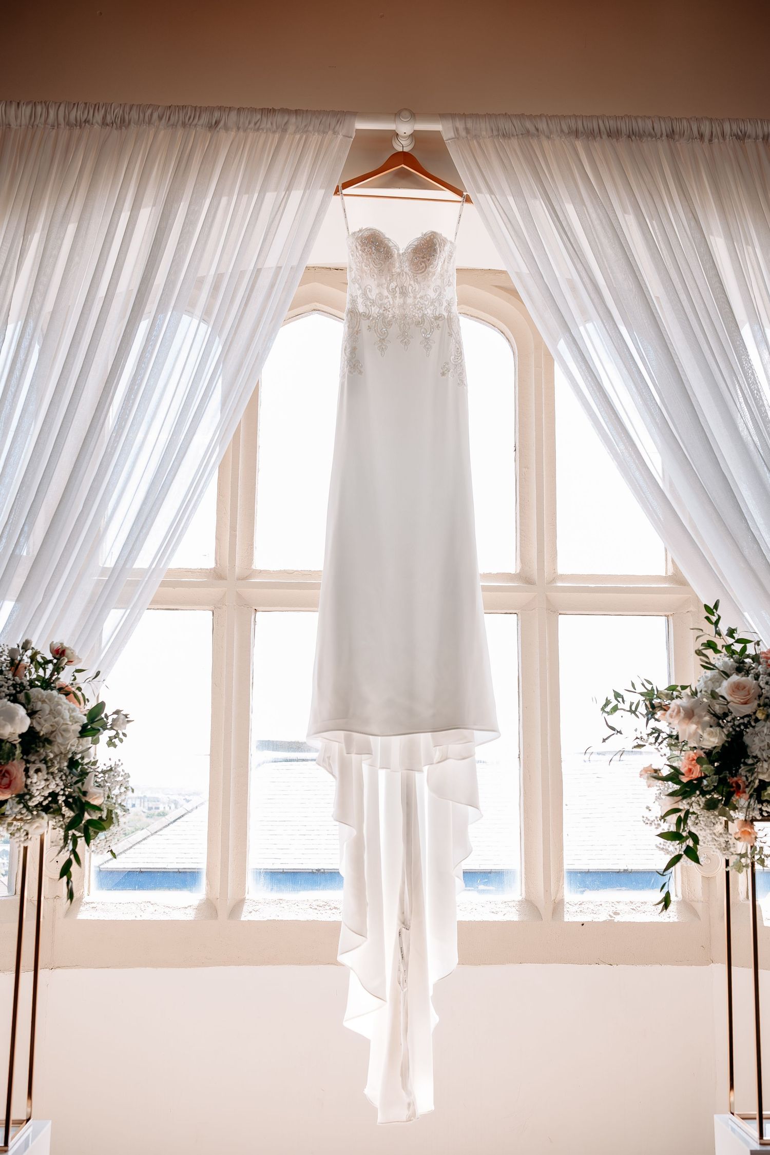Elegant white strapless wedding gown with lace bodice hanging in front of arched window with floral arrangements.