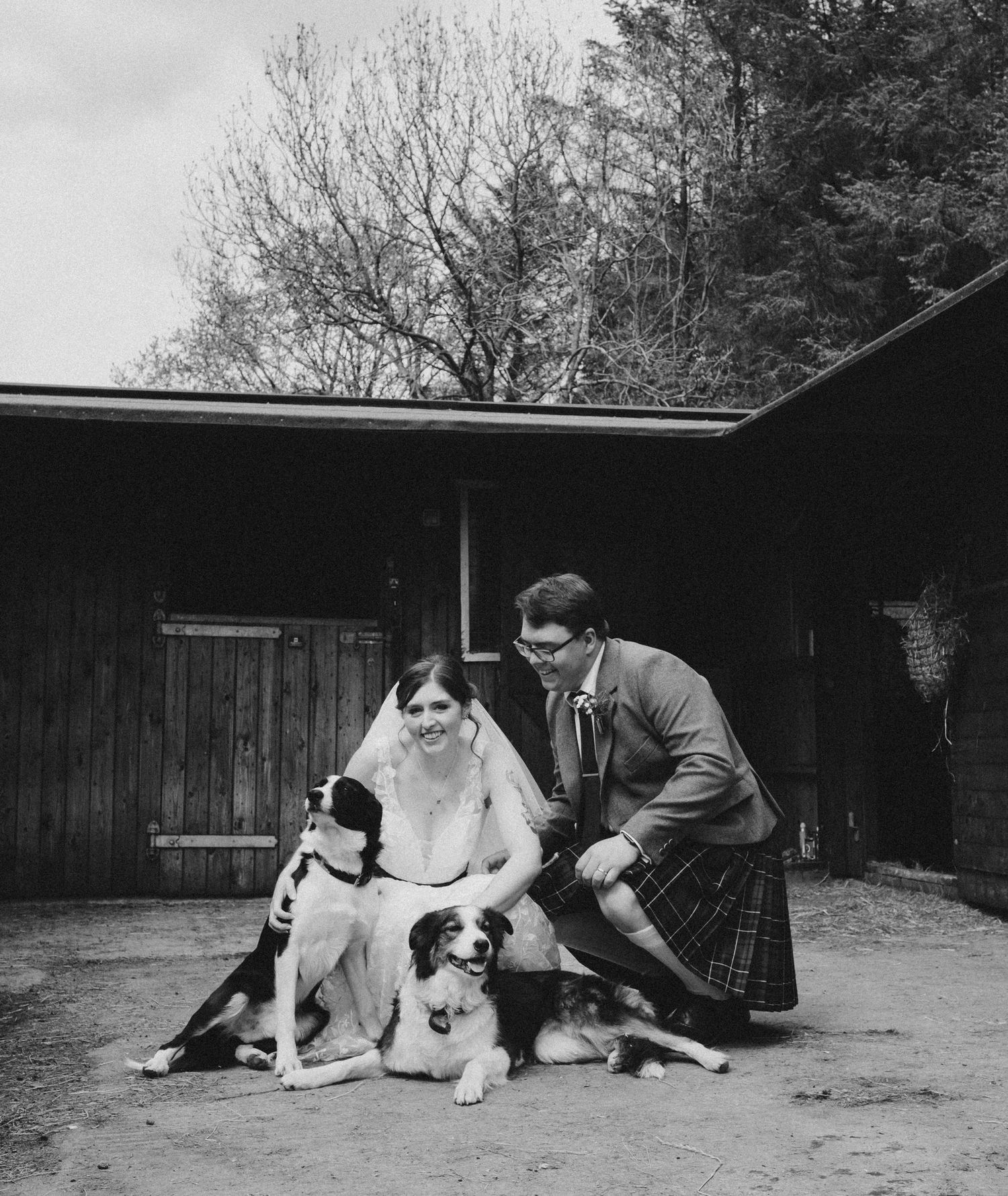 Black and white wedding photo of bride and groom in kilt posing with two dogs outside rustic barn.