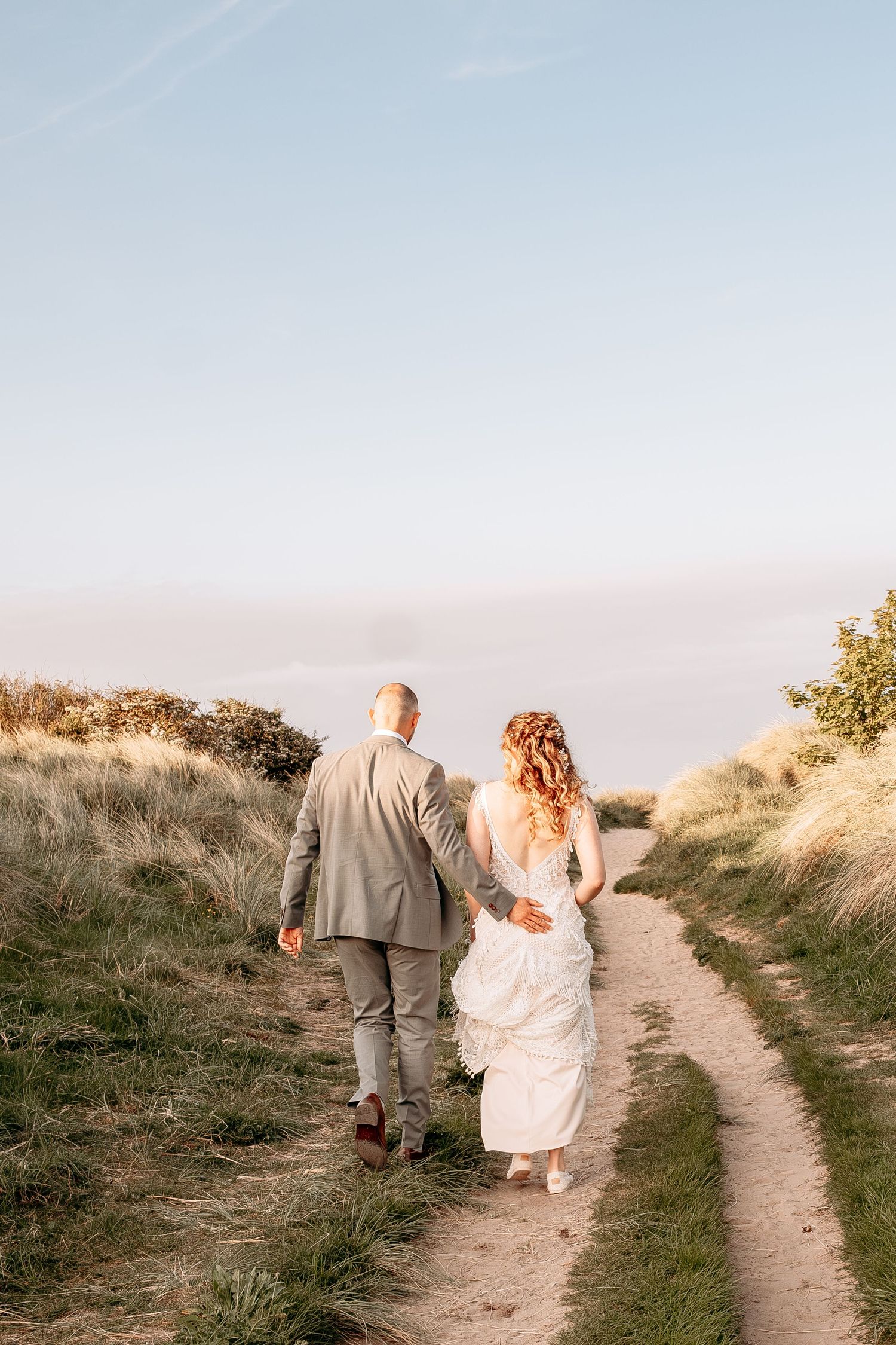Bride and groom walking hand in hand down a sandy path through green dunes at golden hour.
