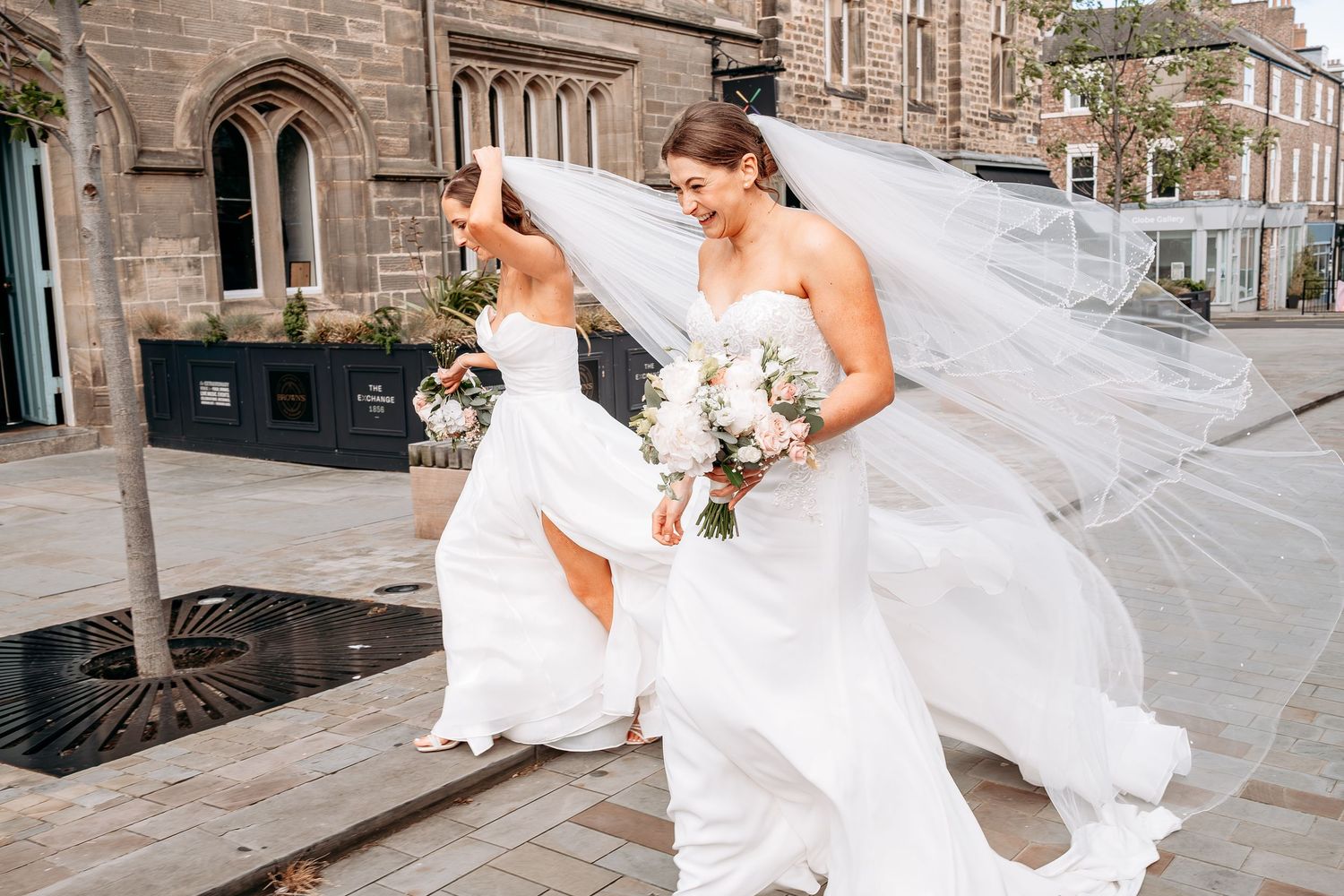 Two brides in white gowns with flowing veils walk outdoors holding bouquets on their wedding day.