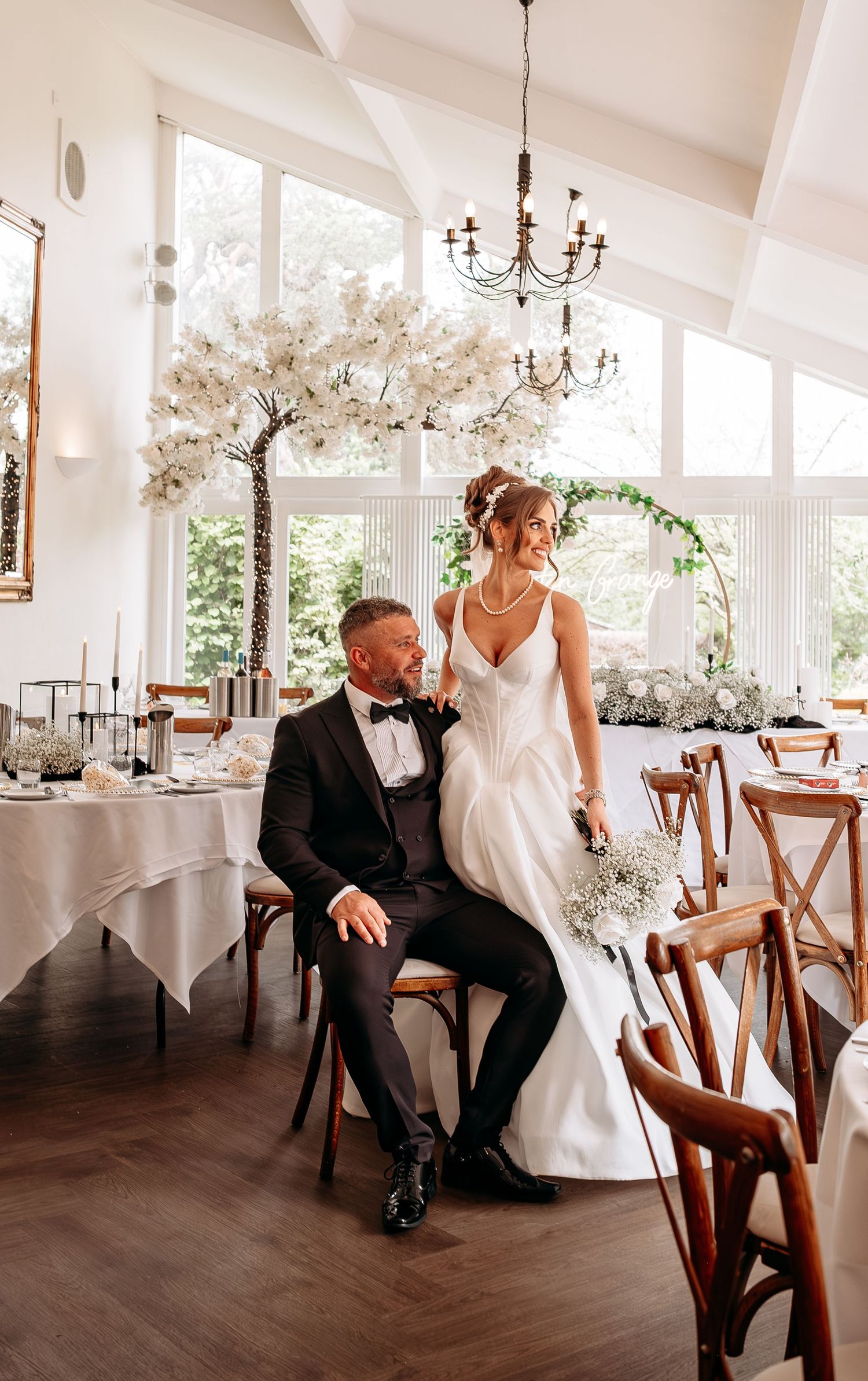 Bride and groom pose elegantly in a bright wedding venue with white floral decor and chandelier.