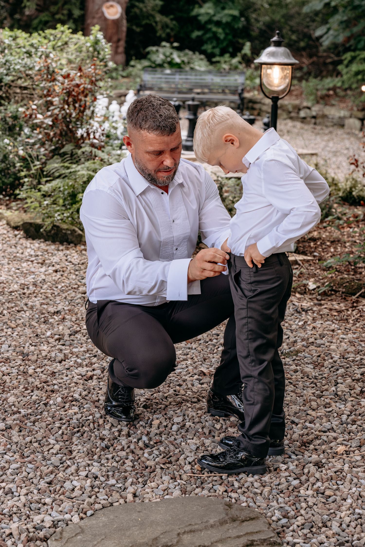 Father in white shirt helps young blonde boy adjust suit on gravel path in garden setting.