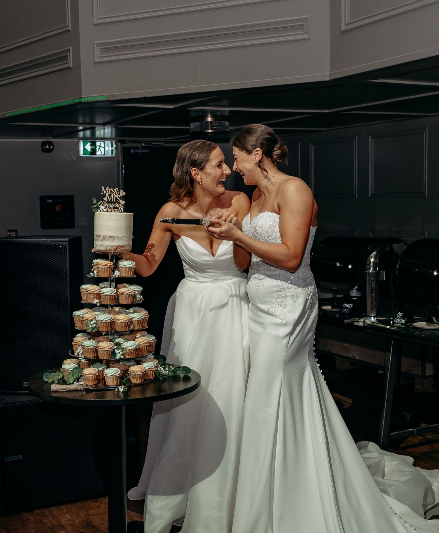 Two brides in white gowns cut their wedding cake together, smiling and touching foreheads at elegant venue.