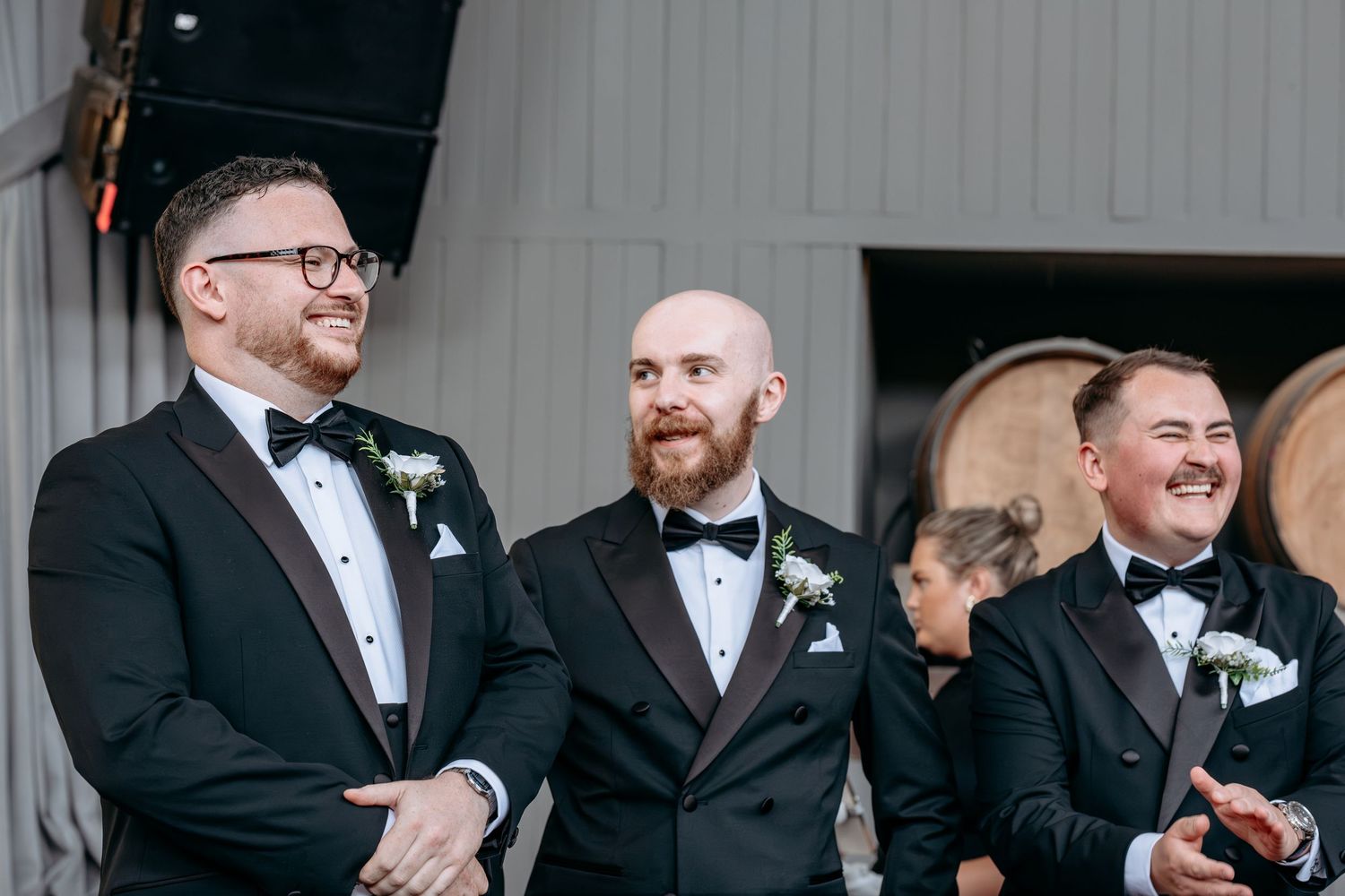 Three groomsmen in black tuxedos with bow ties and boutonnieres smiling at a wedding ceremony.