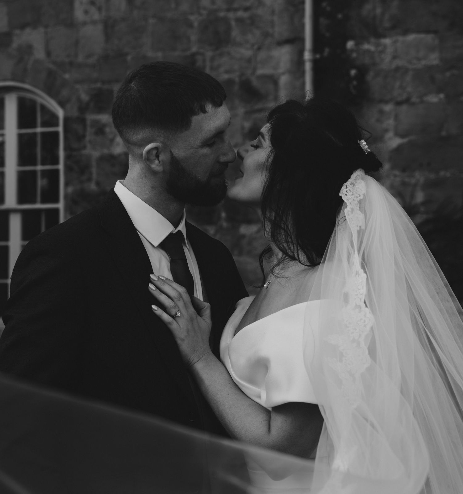 Black and white photo of a bride and groom sharing a romantic kiss on their wedding day near a stone wall.