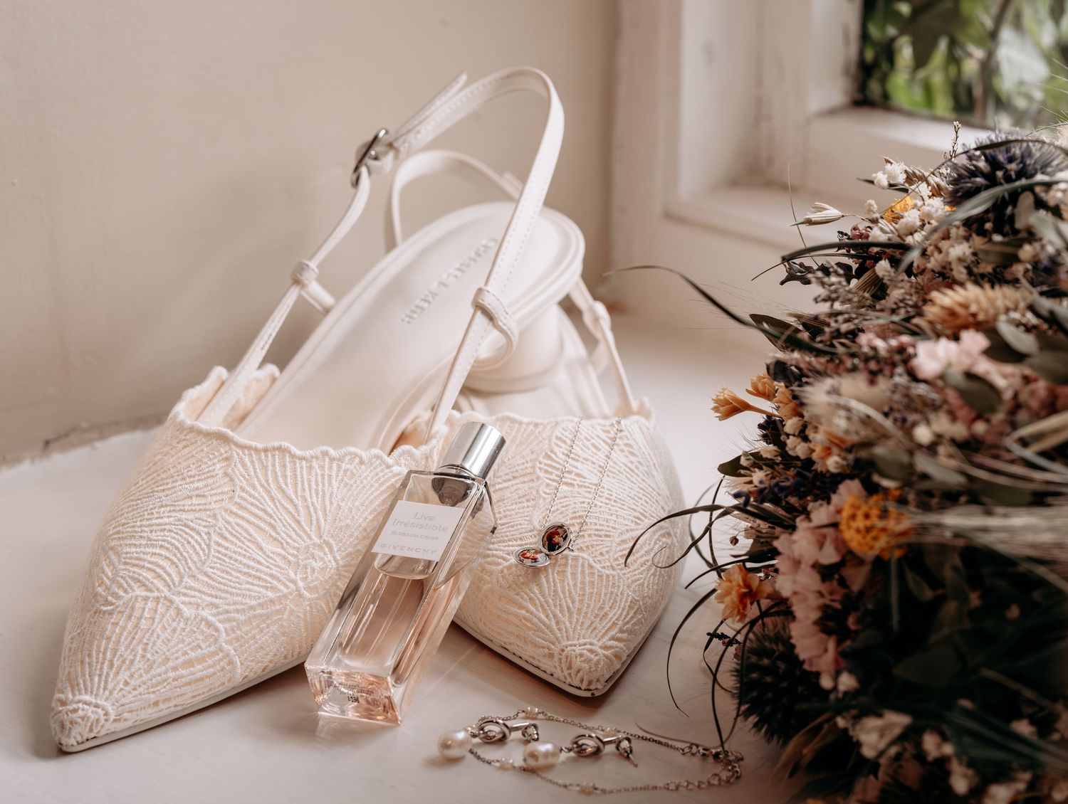 Elegant bridal flat shoes with lace detail, perfume bottle, and dried floral bouquet arranged on a white surface.