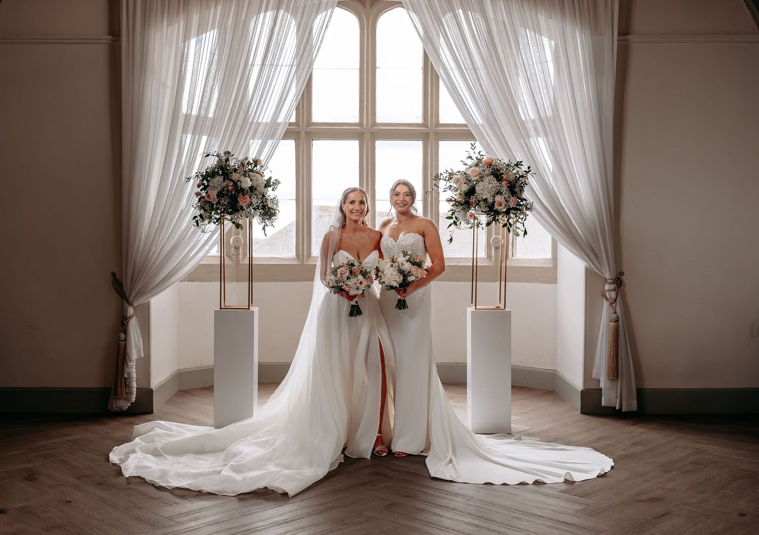 Two brides in elegant white wedding gowns holding bouquets stand before draped windows with floral arrangements.