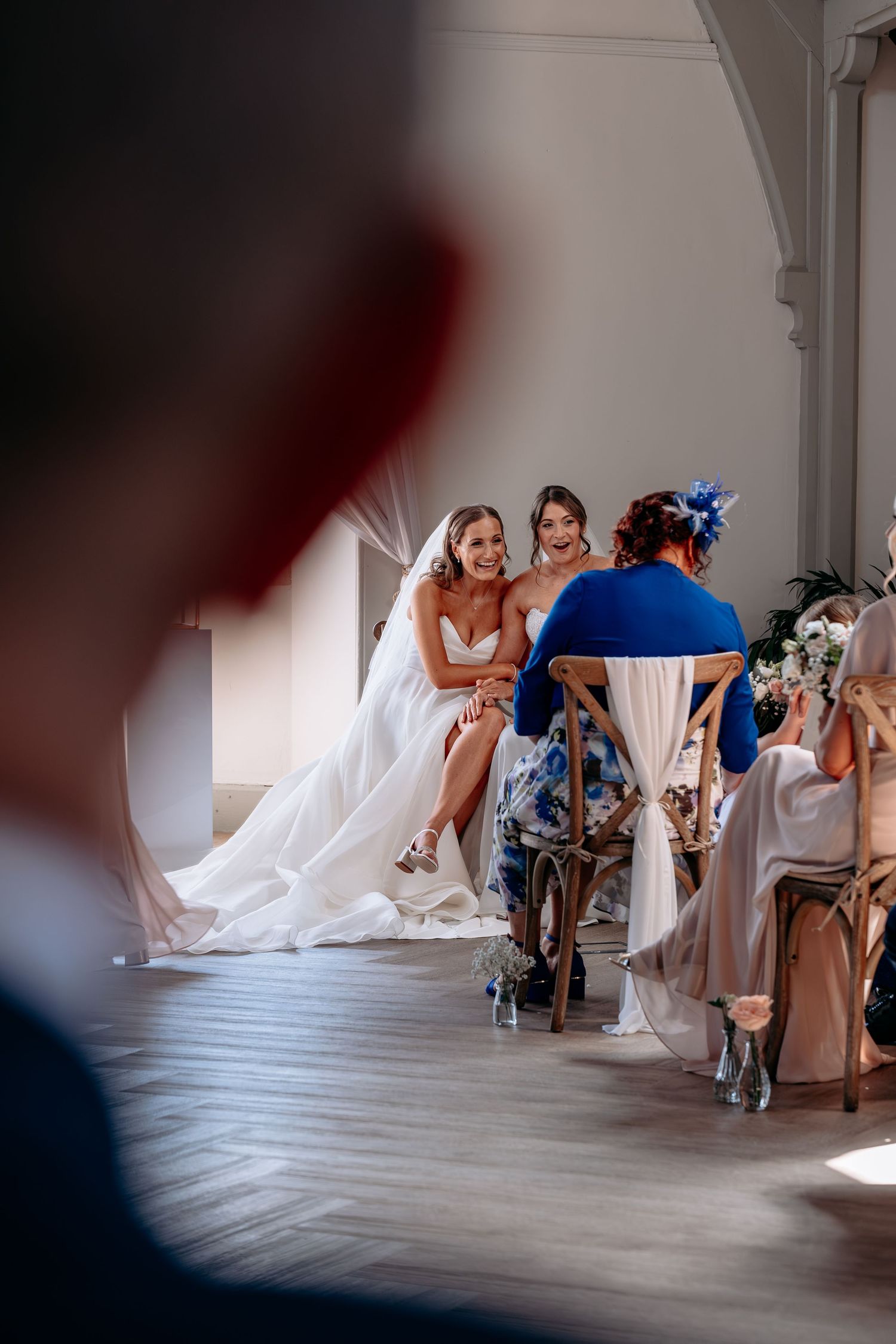 Bride in white gown laughing with bridesmaid and guests seated at wedding reception table in elegant venue.