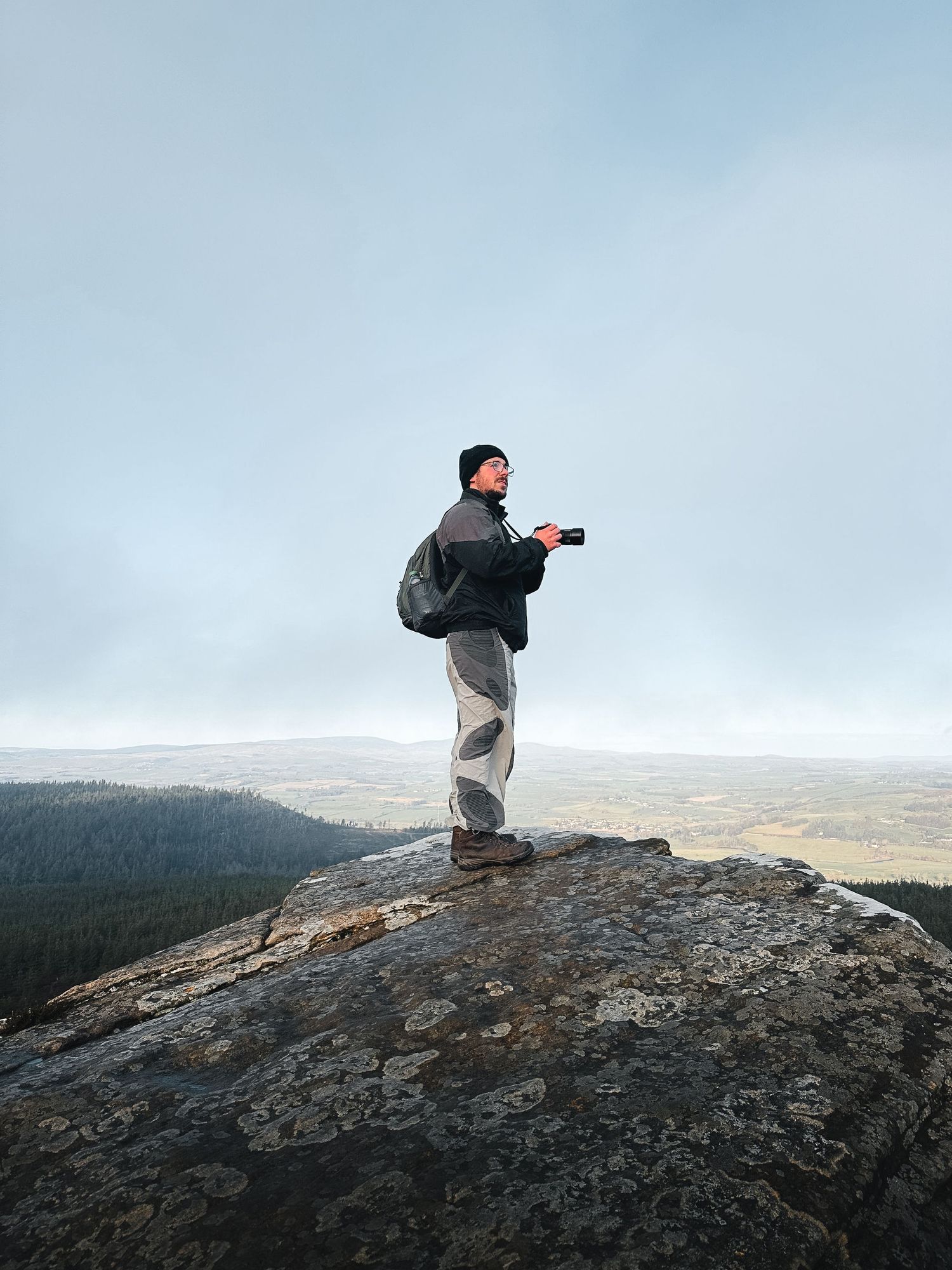 Hiker standing on rocky summit with camera, overlooking vast misty valley and forest landscape under overcast sky.