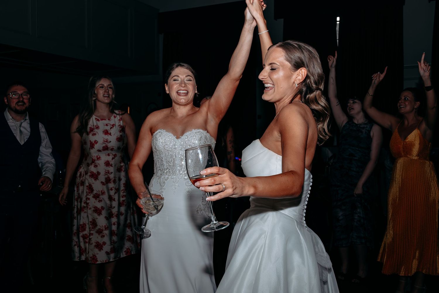 Two brides in white gowns dancing joyfully at their wedding reception, holding champagne glasses with guests celebrating behind them.