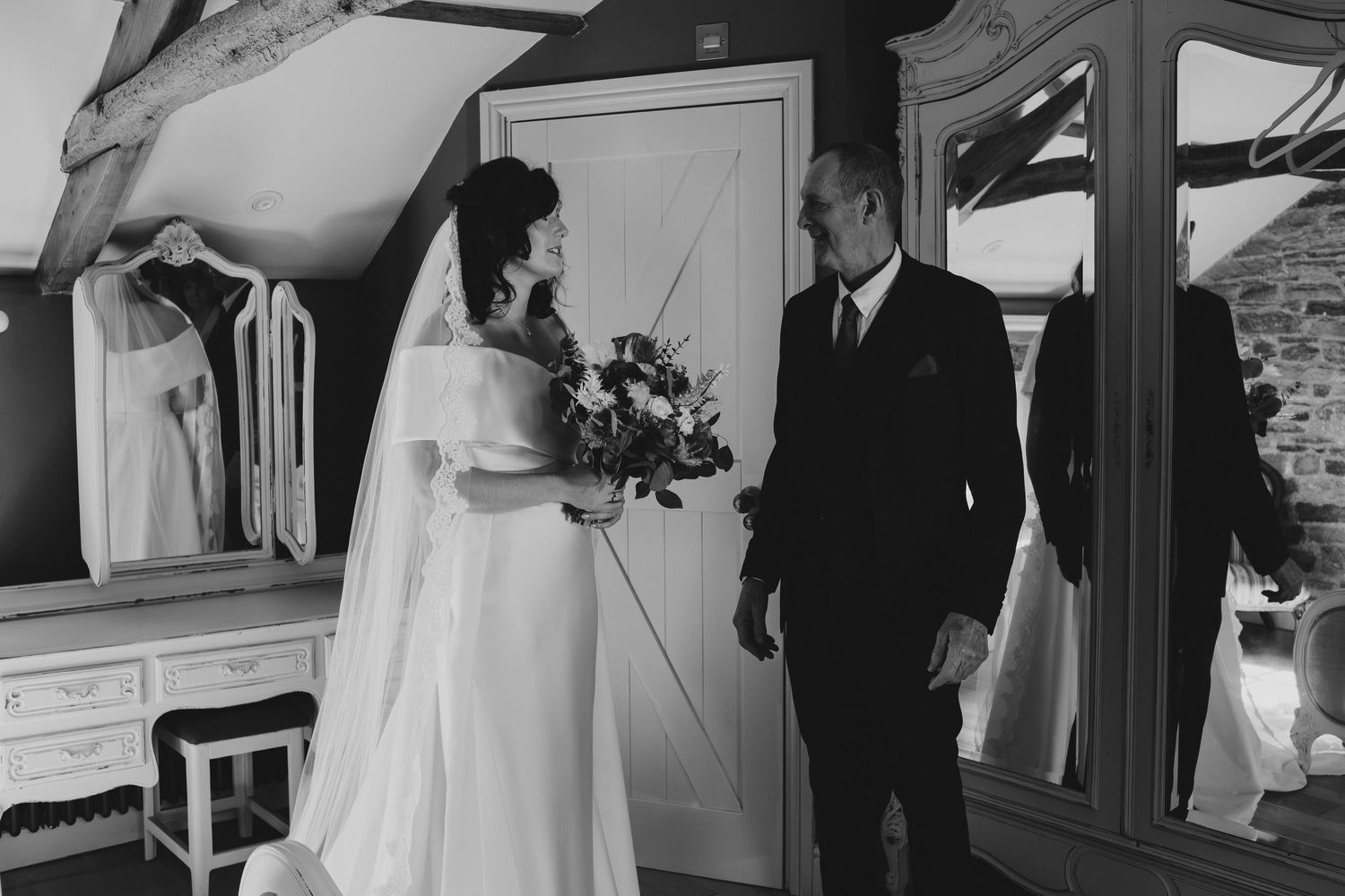 Black and white photo of a bride holding a bouquet standing with a man in a suit in a rustic wedding venue.