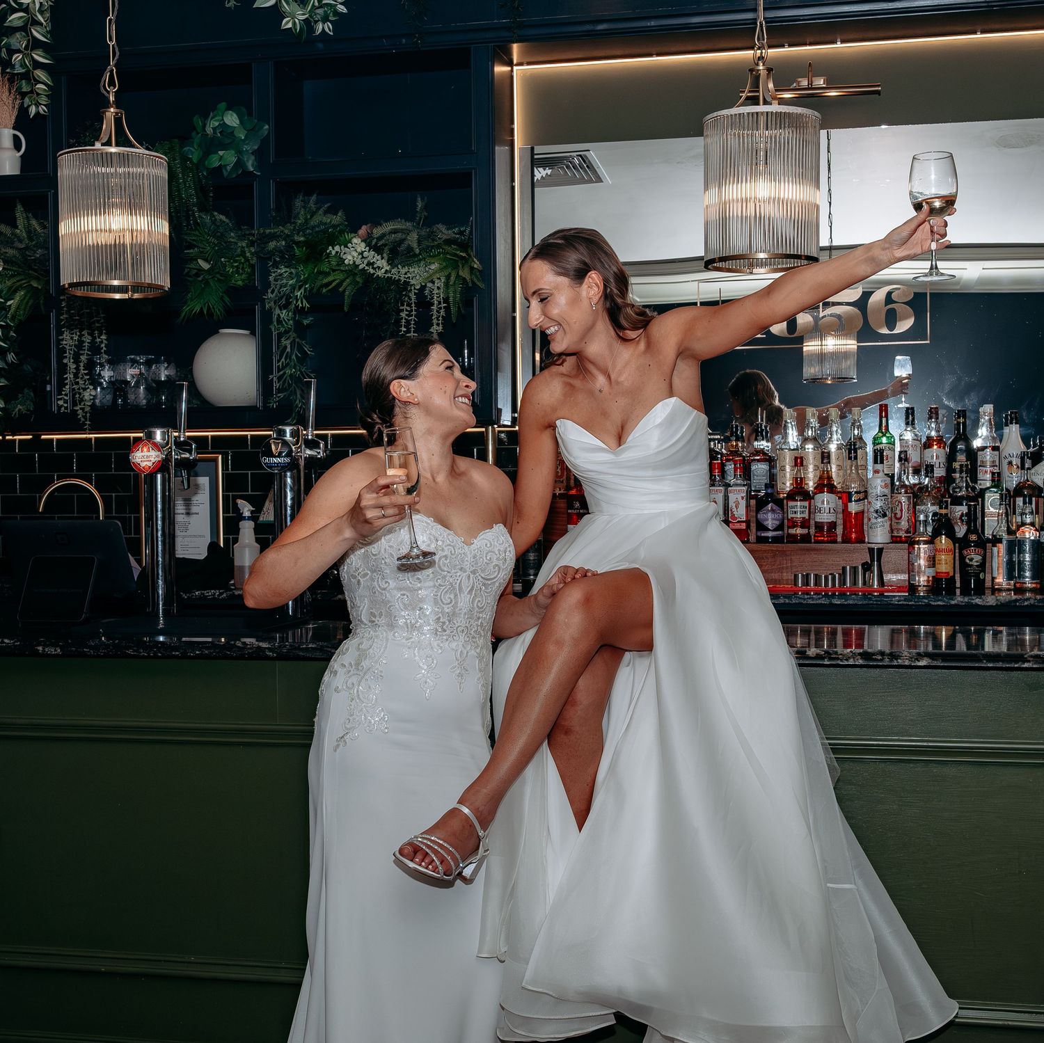 Two brides in white wedding gowns celebrate with wine glasses at an elegant bar with pendant lights.
