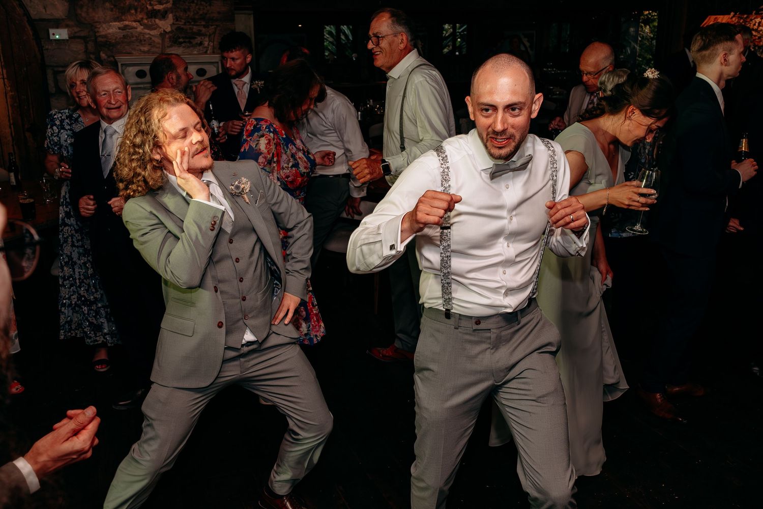 Two men in matching grey suits and suspenders dance energetically at a wedding reception.