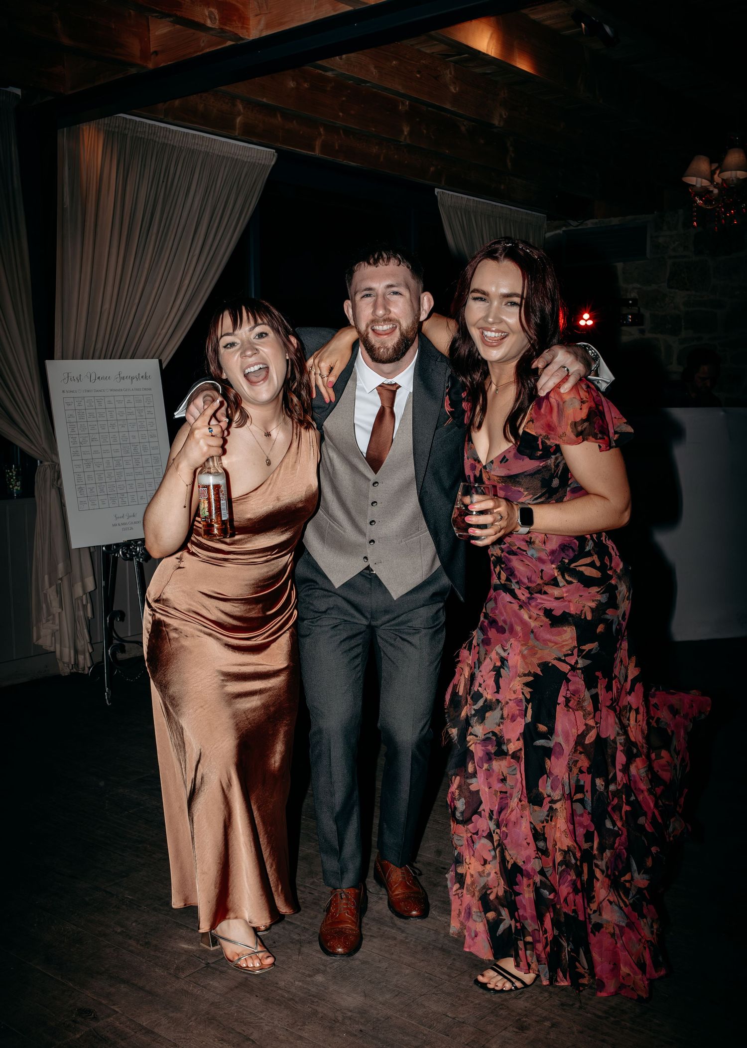 Three friends pose joyfully at a wedding reception, man in gray suit flanked by women in satin and floral gowns.