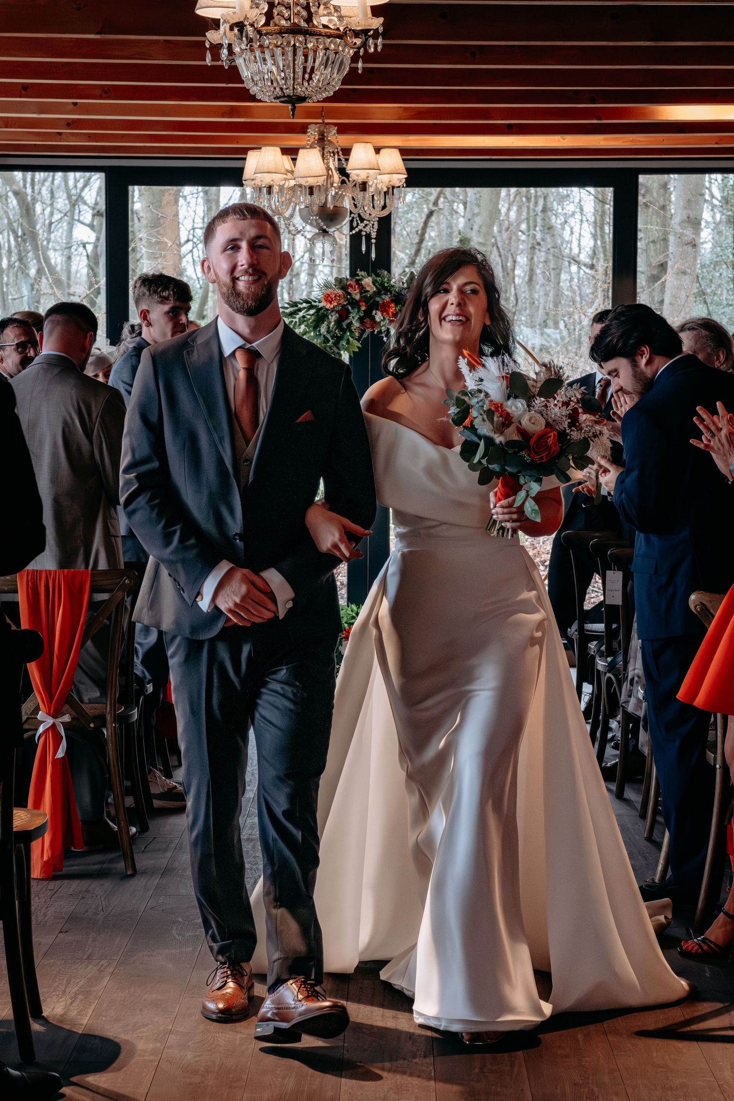 Bride and groom walk down the aisle smiling after wedding ceremony in elegant wood-paneled venue with chandelier.