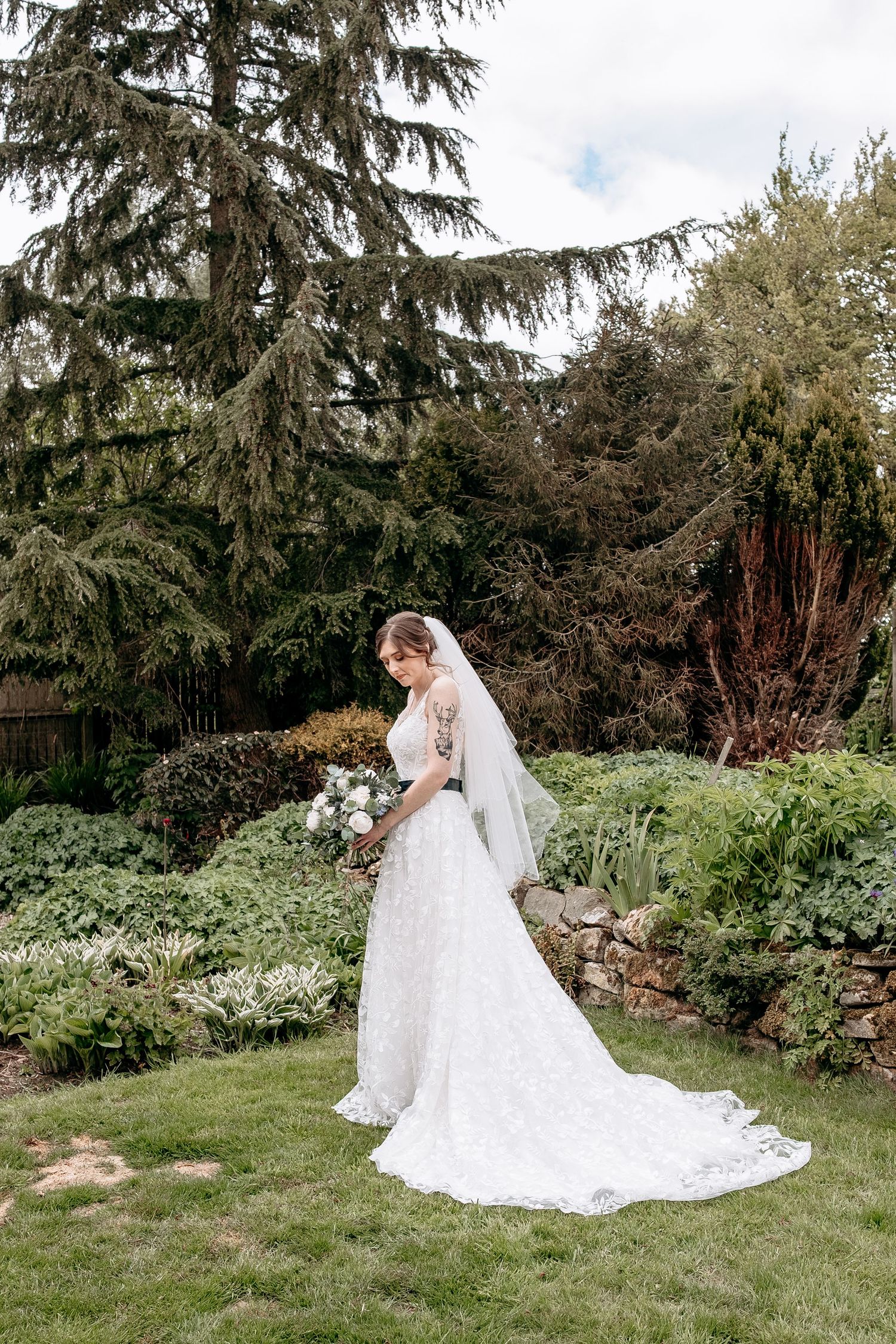 Bride in lace wedding gown with veil holding bouquet in lush garden setting with evergreen trees.