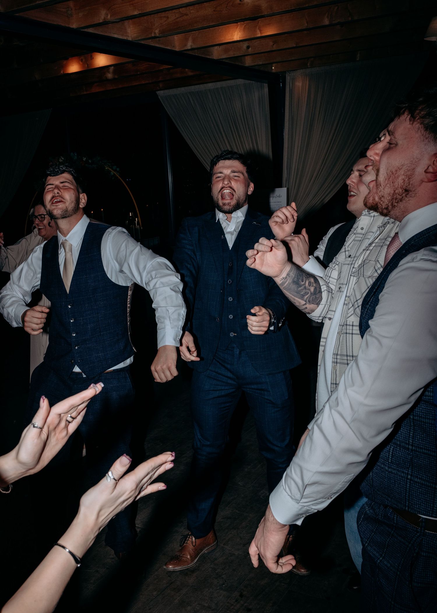 Groomsmen dancing energetically at a wedding reception in navy suits, celebrating with guests in a dimly lit venue.