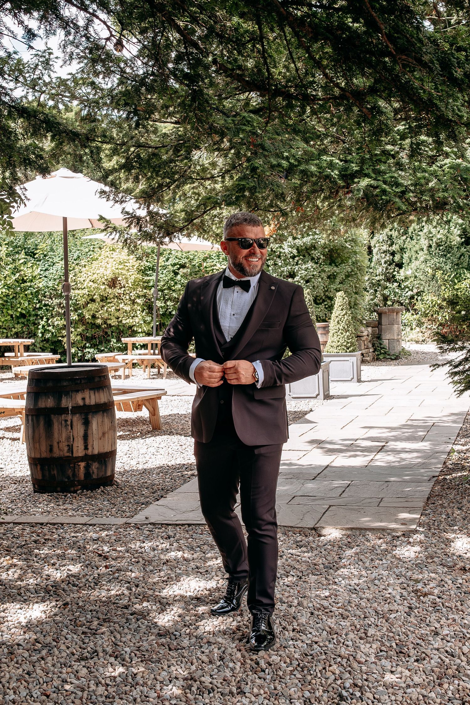 Groom in dark tuxedo with bow tie and sunglasses buttoning jacket in a lush garden setting with wooden barrel.