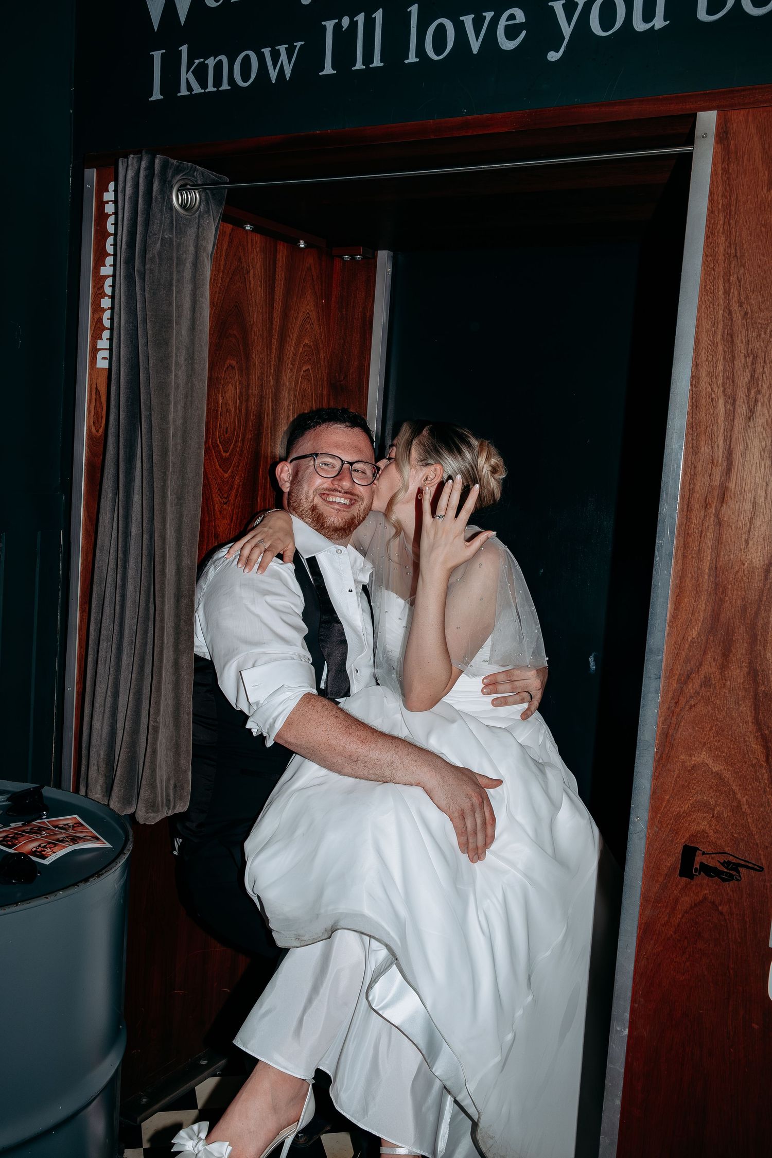A joyful bride and groom share a kiss in a photo booth, with the groom smiling while seated and the bride in a white gown.