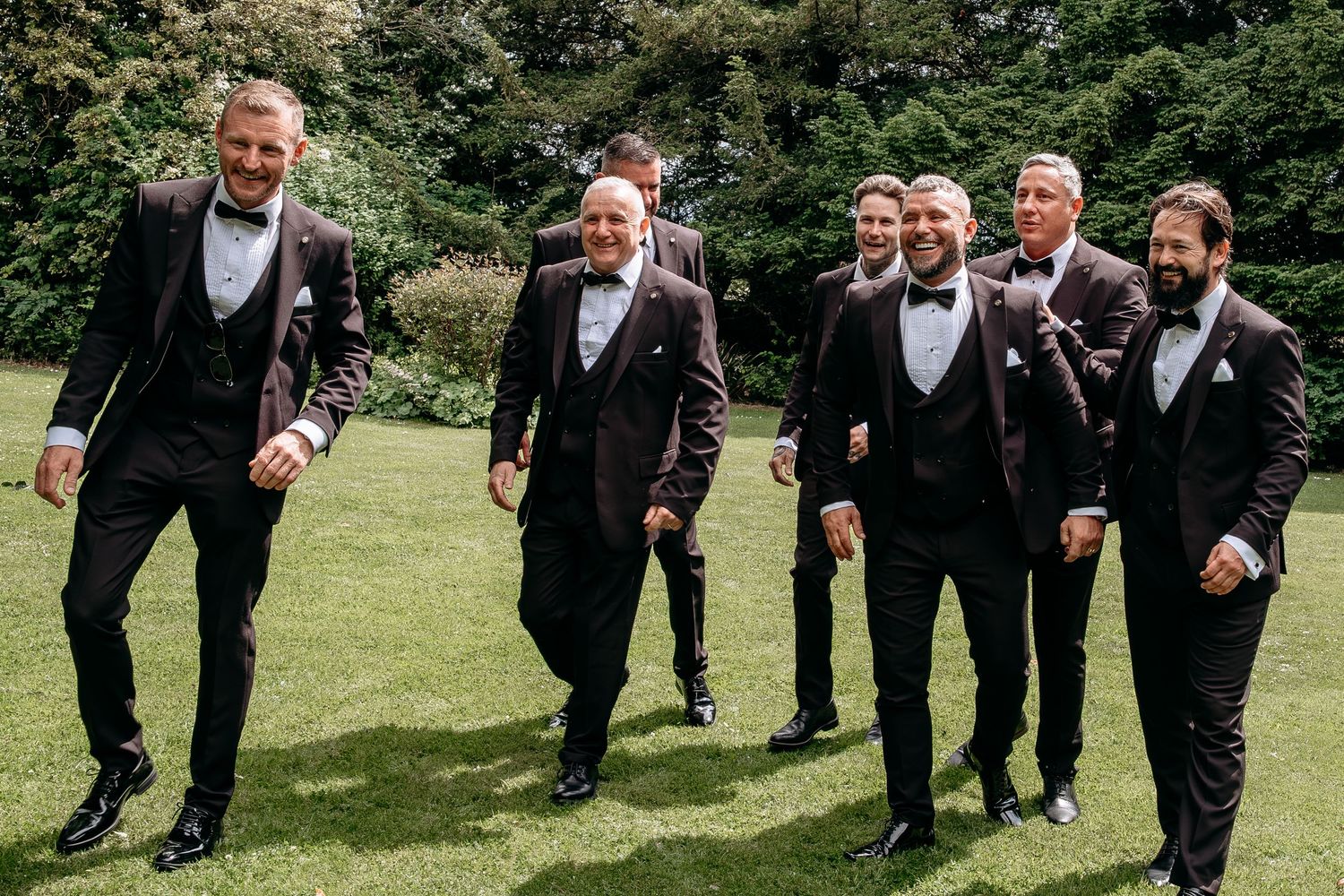 Six groomsmen in black tuxedos with bow ties walking and laughing together on green lawn at outdoor wedding.