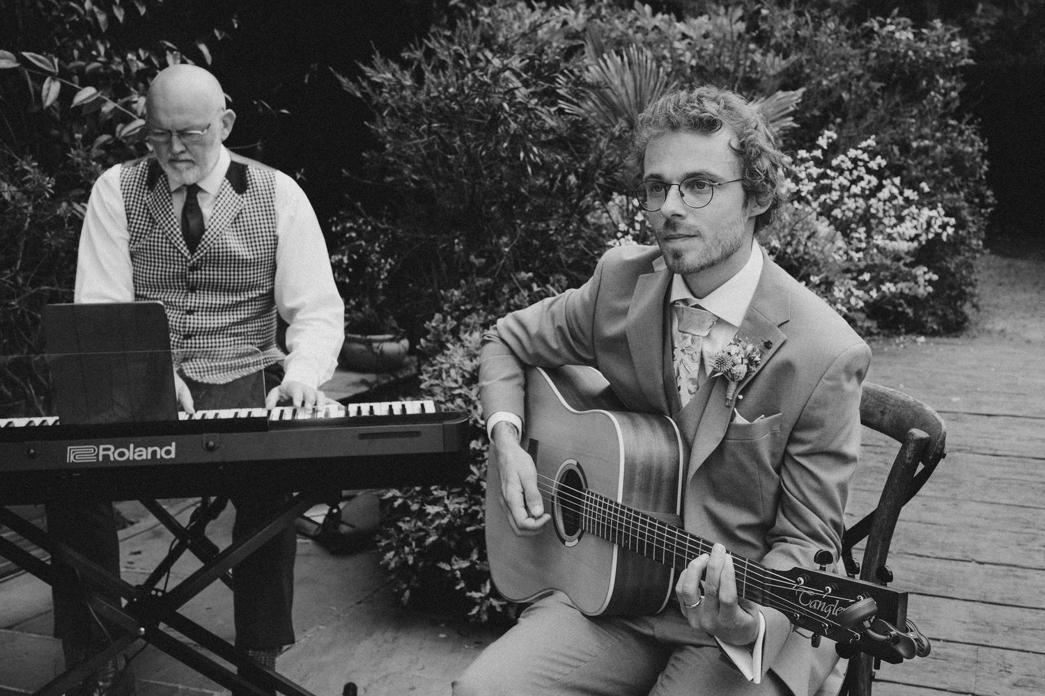 Two musicians perform outdoors, one playing a Roland keyboard and another seated with an acoustic guitar, in black and white.