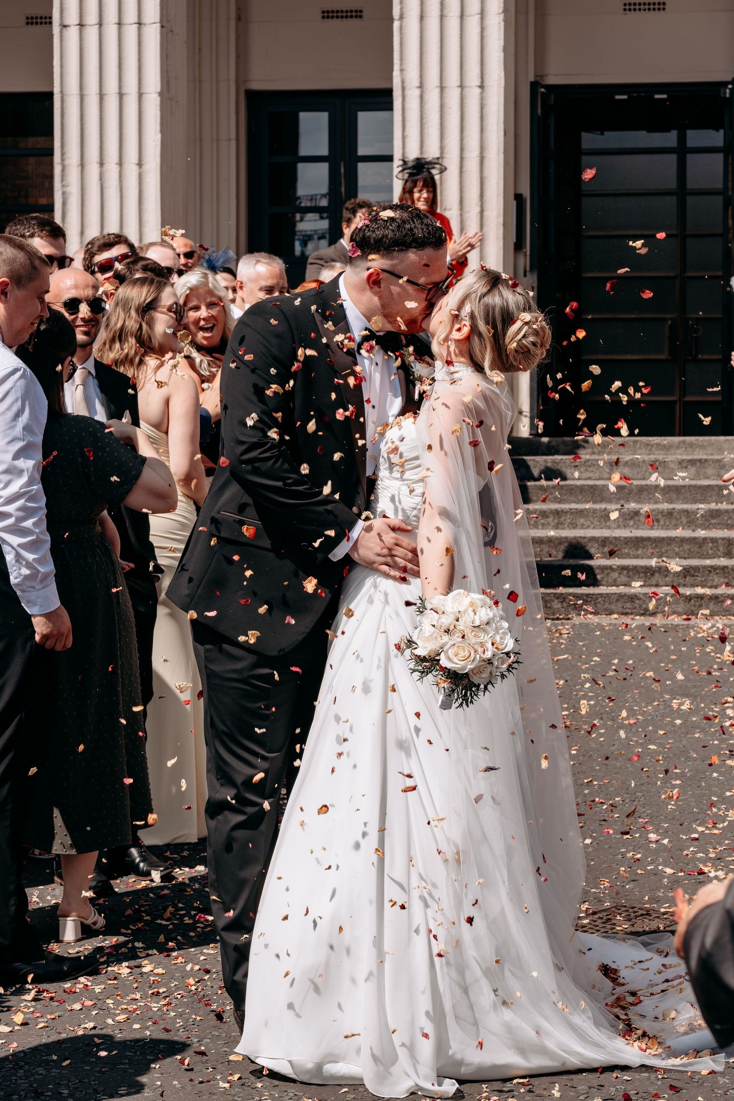 Bride and groom kissing outside church surrounded by guests throwing colorful confetti on their wedding day.
