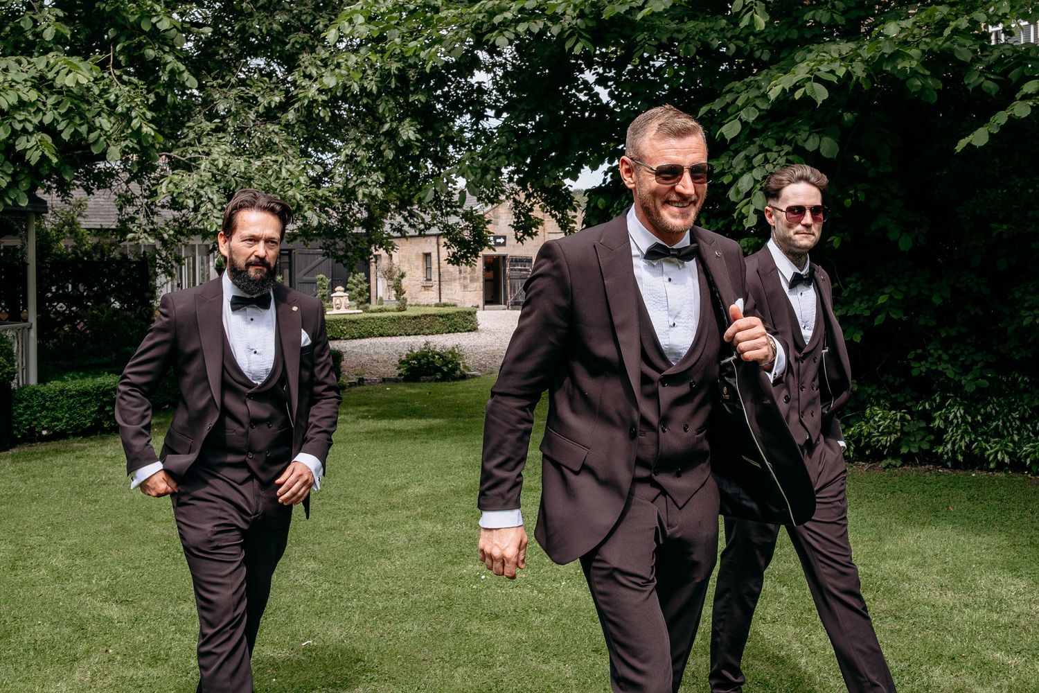 Three groomsmen in dark brown suits with bow ties walking confidently across a green lawn at a wedding.