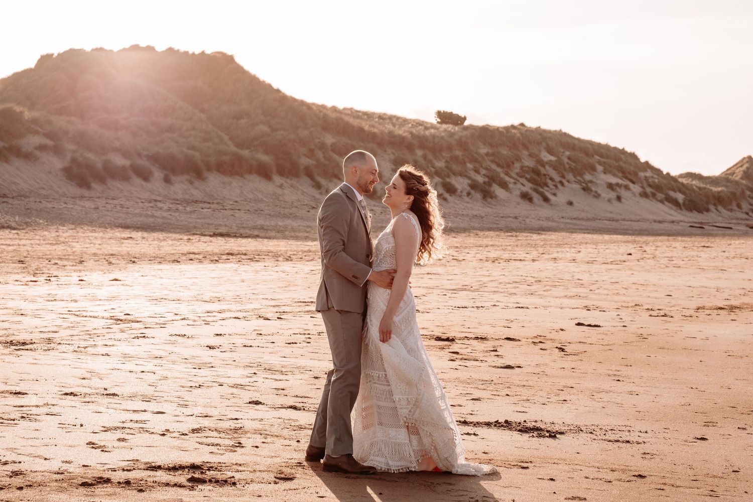 Bride and groom share a romantic embrace on a sandy beach at golden sunset with sand dunes in the background.
